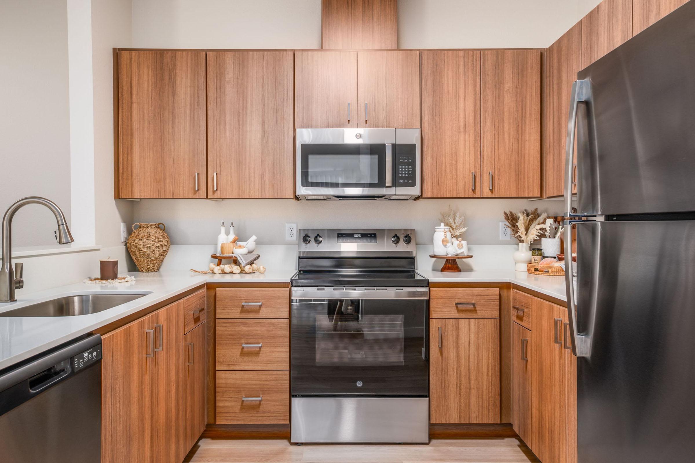 A modern kitchen featuring warm wooden cabinetry, stainless steel appliances, and a sleek countertop. The space includes a microwave above an oven, a double sink, and decorative elements like small vases and a basket, creating a cozy and inviting atmosphere.