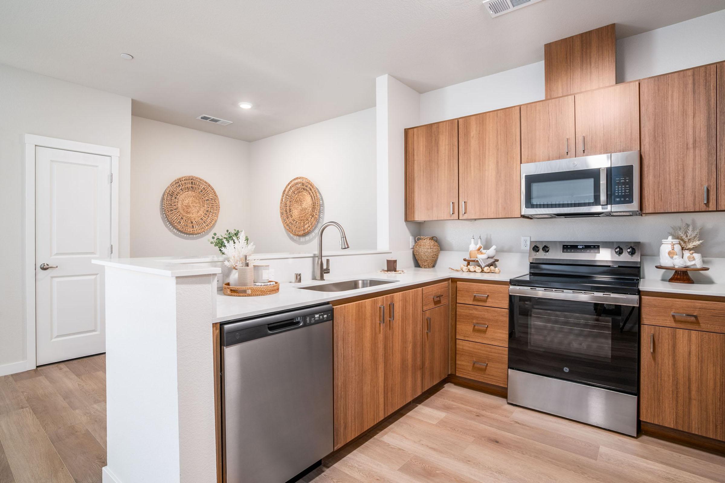 Modern kitchen featuring wooden cabinetry, a stainless steel microwave and oven, a dishwasher, and a double sink with a sleek faucet. The design includes light-colored walls, wood flooring, and decorative woven wall art, creating a warm and inviting atmosphere.