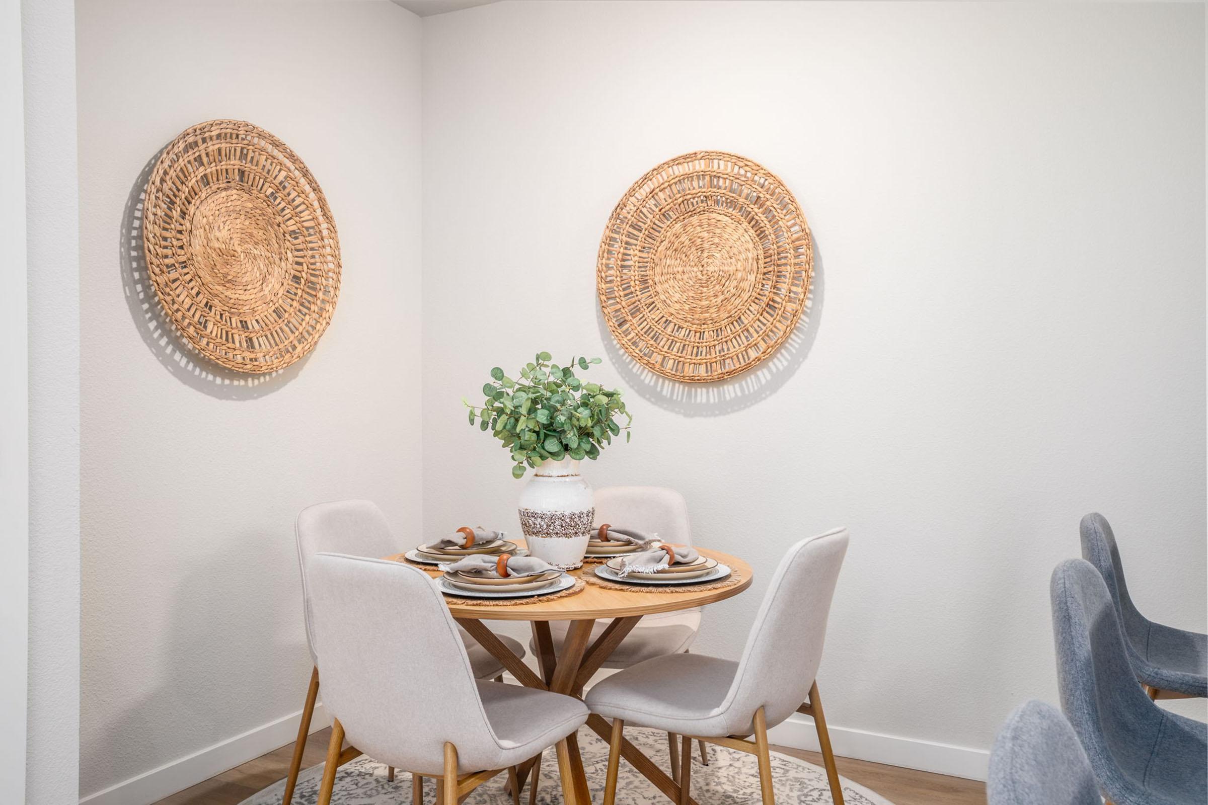 A modern dining area featuring a round wooden table set for four, with decorative plates and cutlery. A white vase with greenery serves as the centerpiece. Two woven wall hangings are displayed on the white walls, complementing the light-colored furniture and wooden floor.