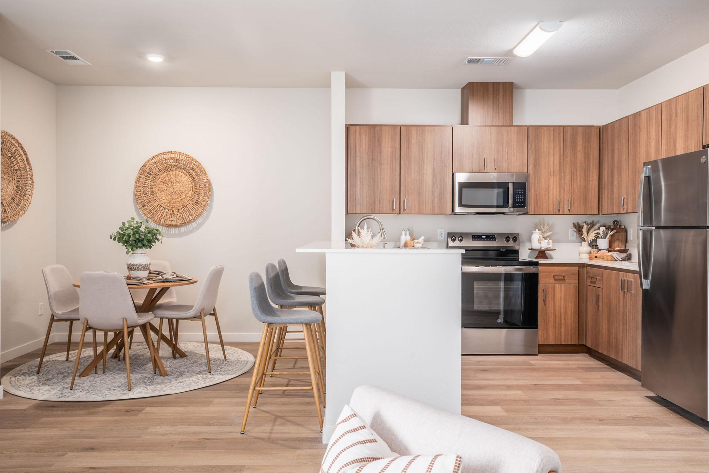Modern kitchen and dining area with light wood cabinetry, stainless steel appliances, and a round dining table set for four. The decor features woven wall art and a cozy seating area with a light-colored armchair, creating a stylish and inviting atmosphere.