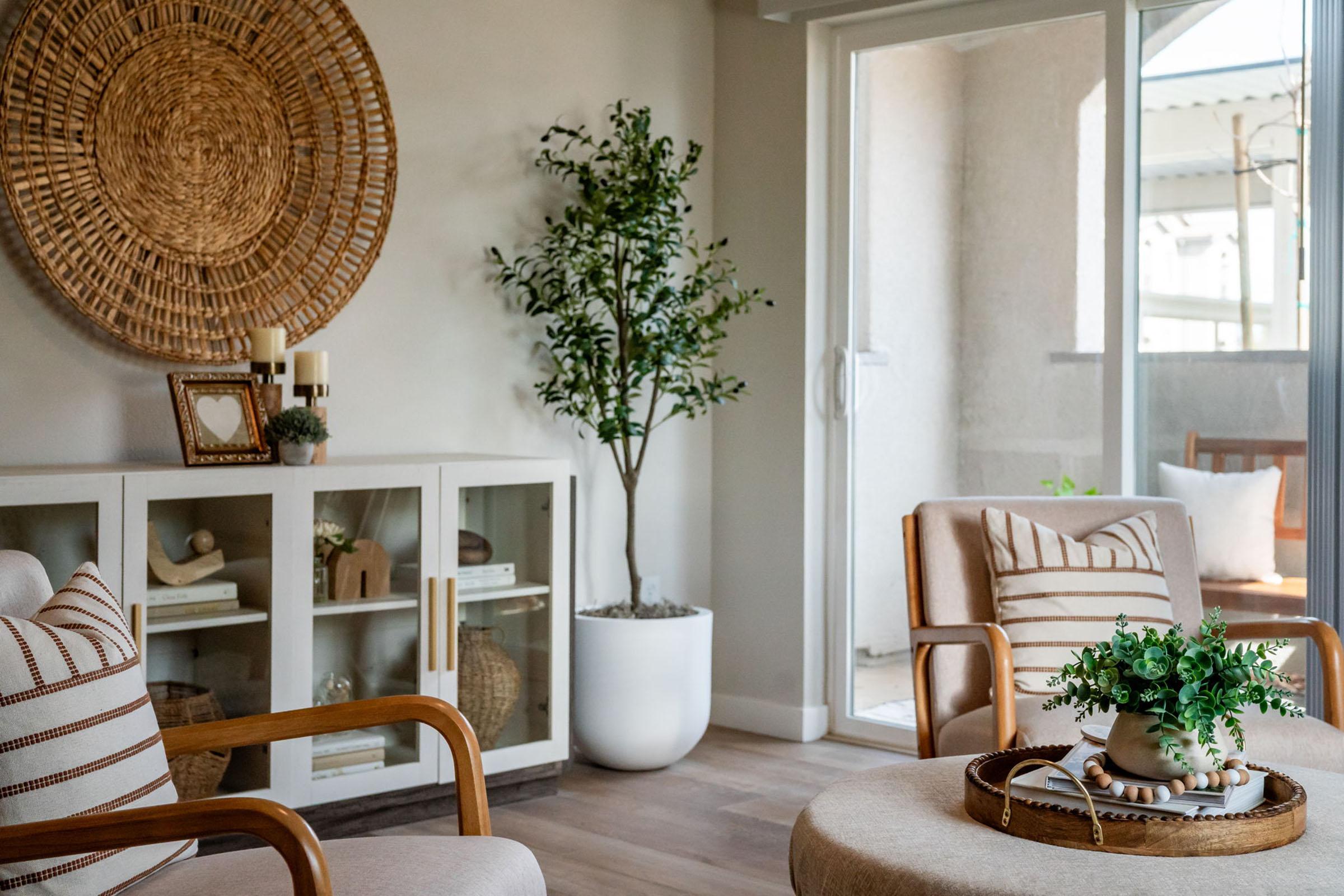A cozy living room featuring two beige armchairs with wooden frames, a round coffee table with a potted plant, and a decorative tray. Behind them, a white cabinet displays decorative items, and a large woven wall art piece hangs above. A potted plant stands next to a glass door leading to a balcony.