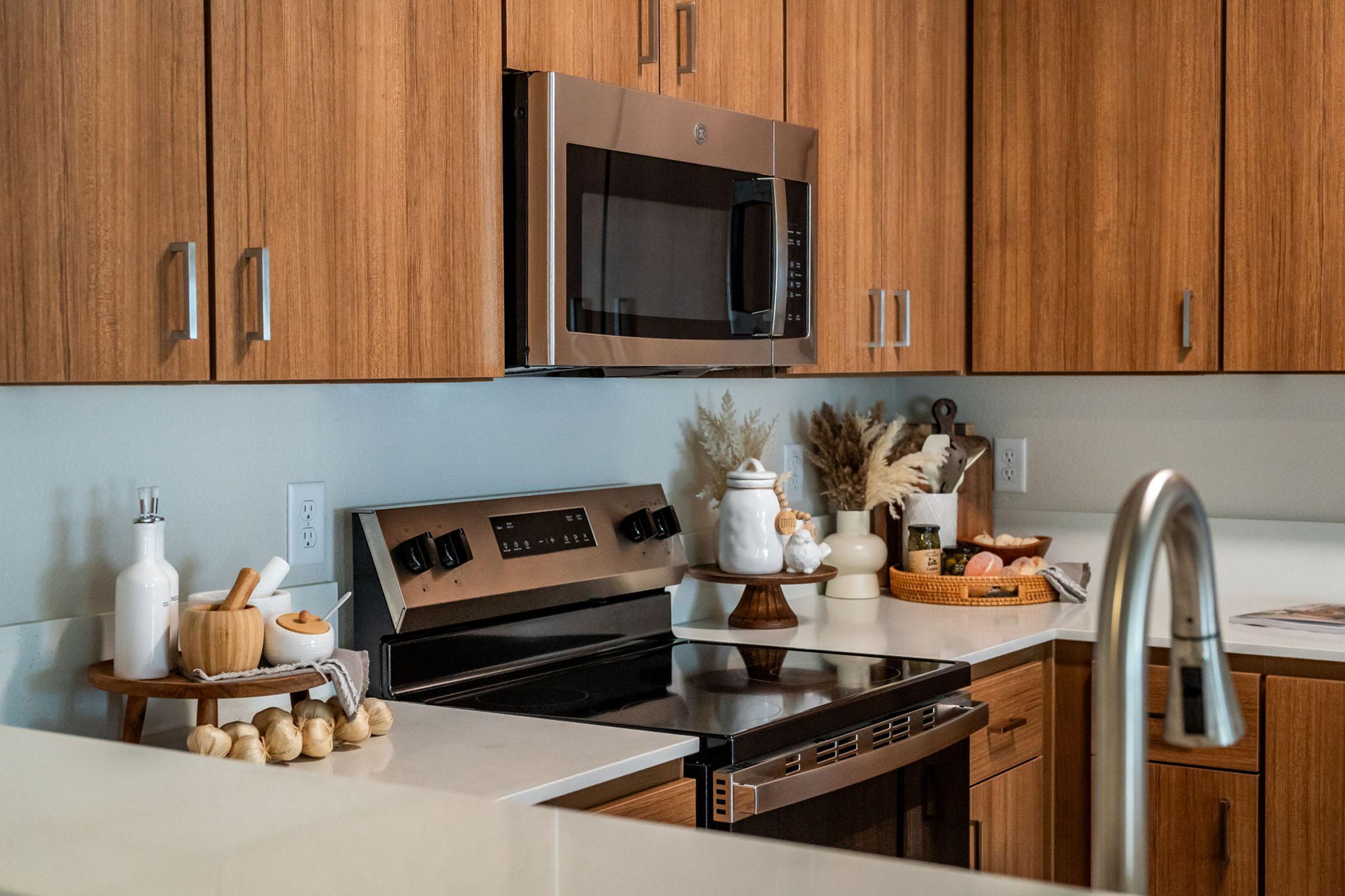 A modern kitchen featuring wooden cabinetry, a stainless steel microwave, and an oven. On the countertop is a white teapot, a decorative bowl, and a wooden utensil holder. Dried plants add a touch of elegance, while a sleek faucet complements the minimalist design.