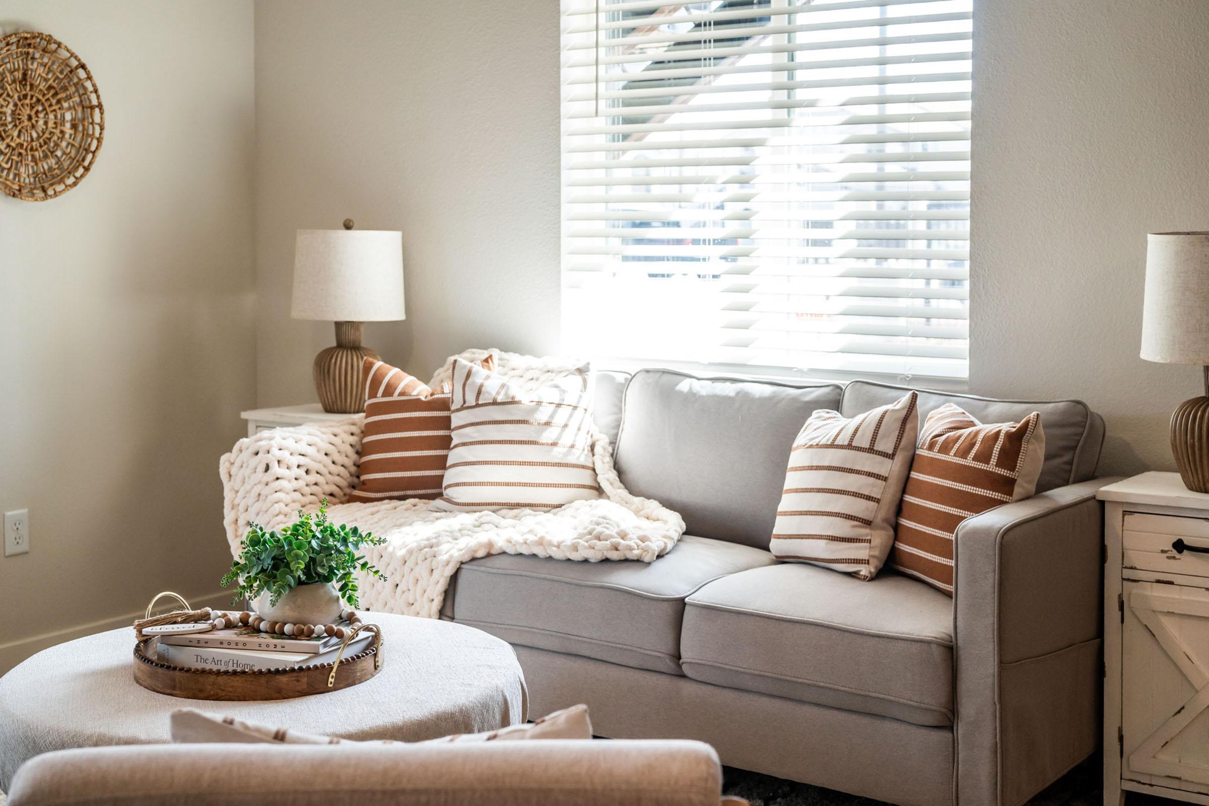 A cozy living room featuring a light gray sofa adorned with striped pillows and a chunky knit blanket. A round coffee table holds decorative items, while a pair of lamps provide soft lighting. Sunlight streams through a window with blinds, creating a warm ambiance.
