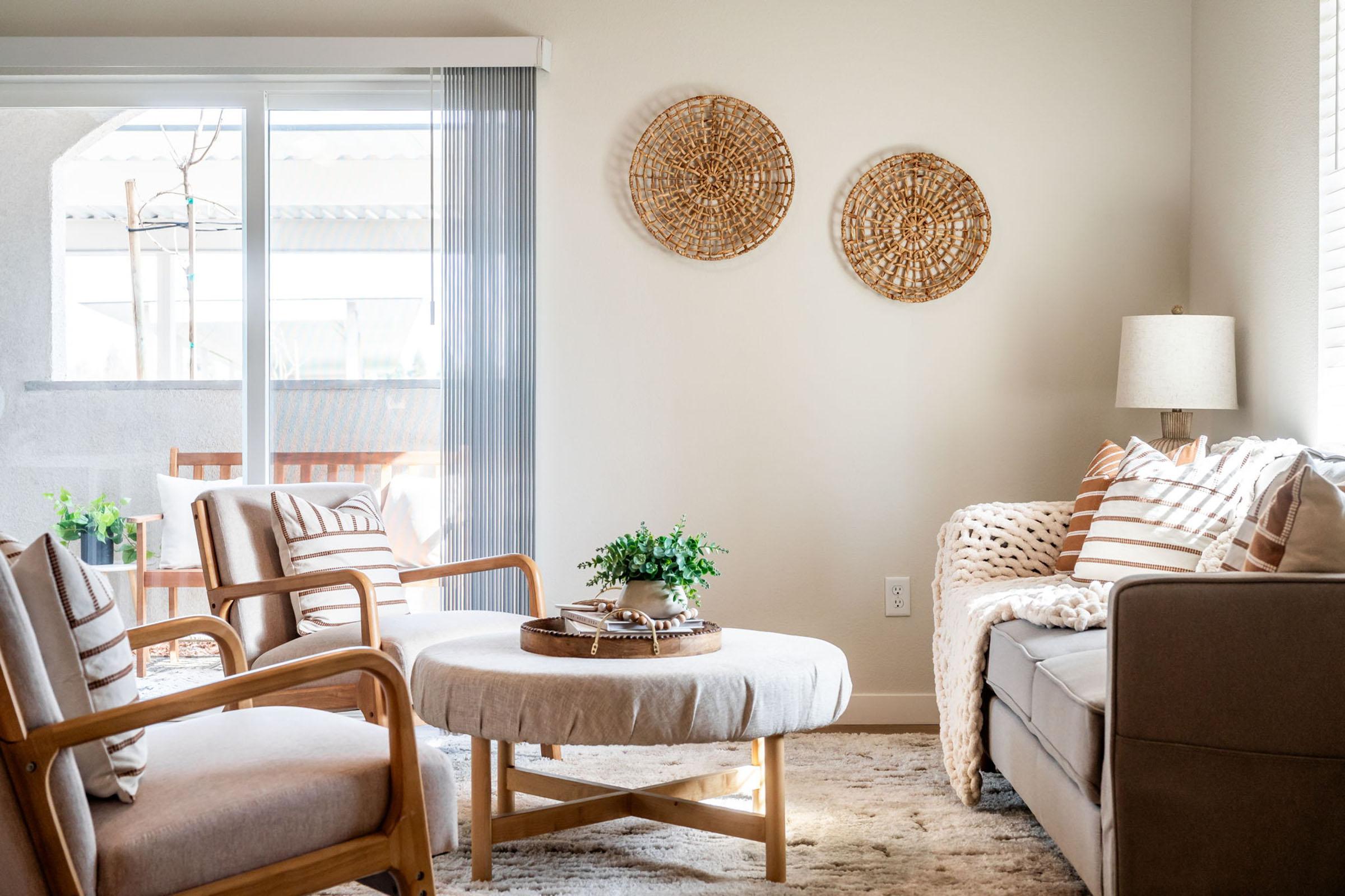 A cozy living room featuring a light-colored sofa with decorative pillows, a round coffee table with a plant, and two wooden armchairs. The space is decorated with woven wall art and has large windows that allow natural light to enter, creating a warm and inviting atmosphere.