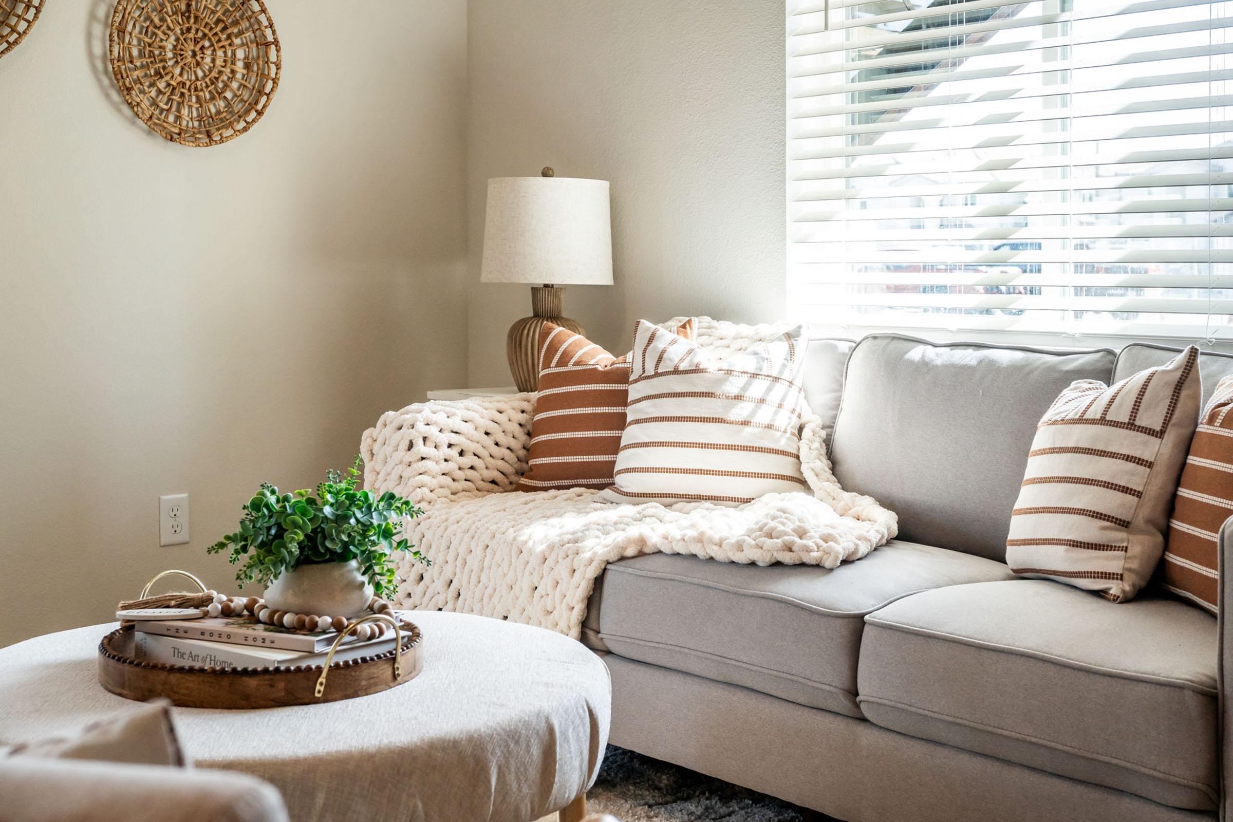 A cozy living room featuring a light gray sectional sofa with striped throw pillows and a chunky knit blanket draped over it. A round coffee table with a decorative tray sits in front, while a lamp and potted plant add warmth to the space. Sunlight filters through white blinds, enhancing the inviting atmosphere.