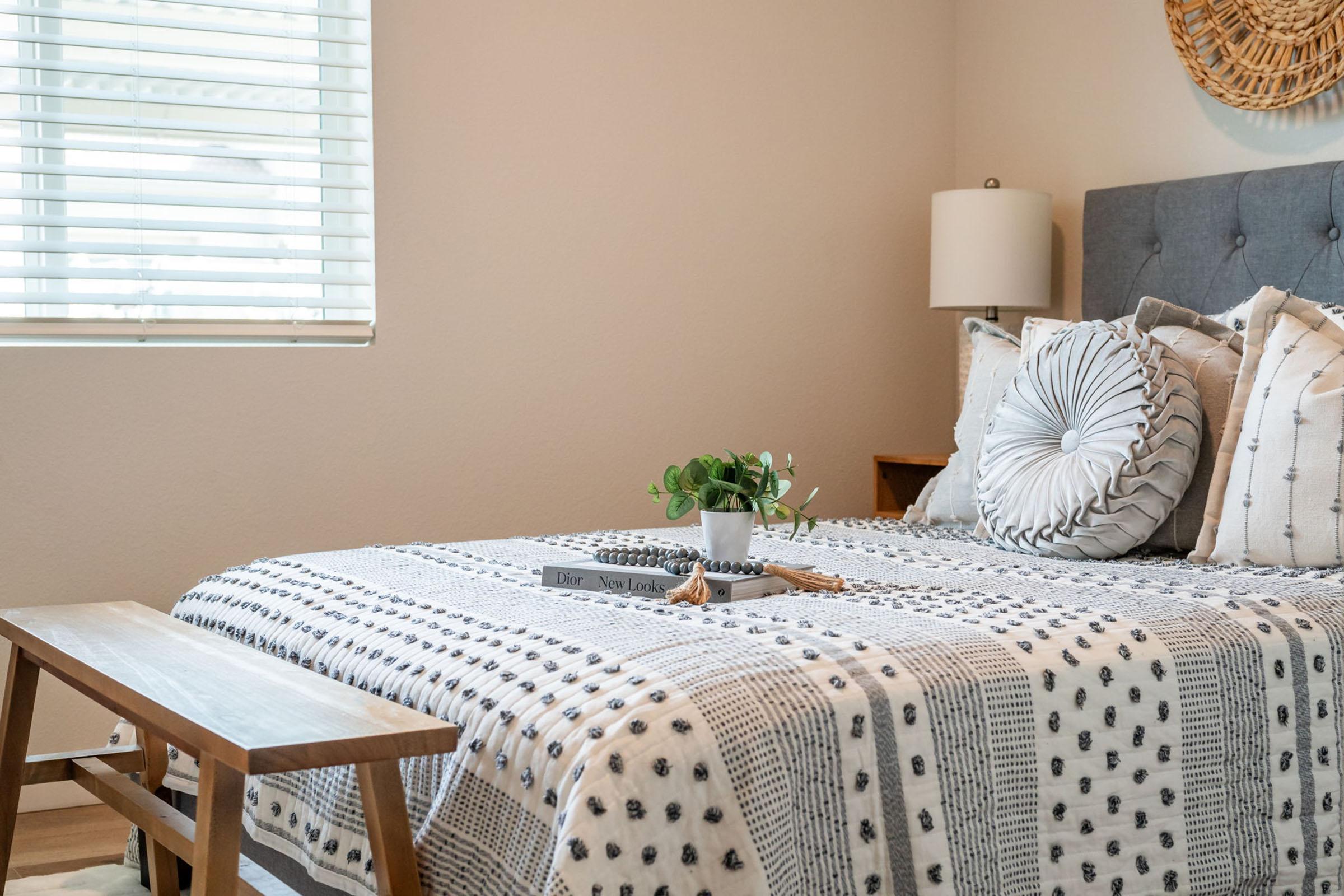 A cozy bedroom featuring a neatly made bed with patterned bedding and decorative pillows. A small plant sits on the nightstand next to a lamp. A wooden bench is at the foot of the bed, and natural light filters through a window with blinds, creating a warm and inviting atmosphere.
