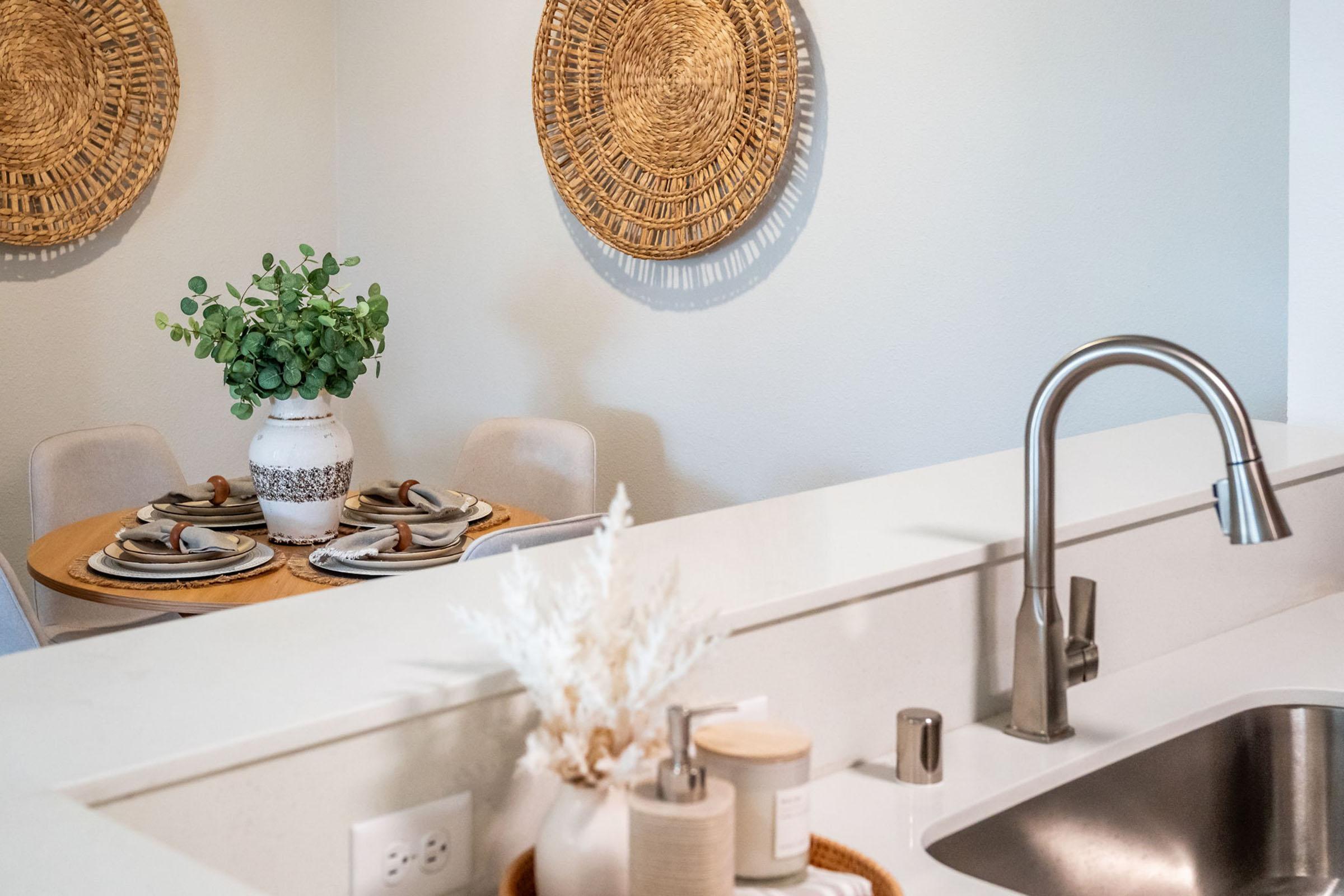 A modern kitchen featuring a sleek sink and countertop. In the background, a dining area with a wooden table set for two, adorned with plates and cutlery. A decorative vase with greenery and woven wall art adds a touch of elegance to the space.