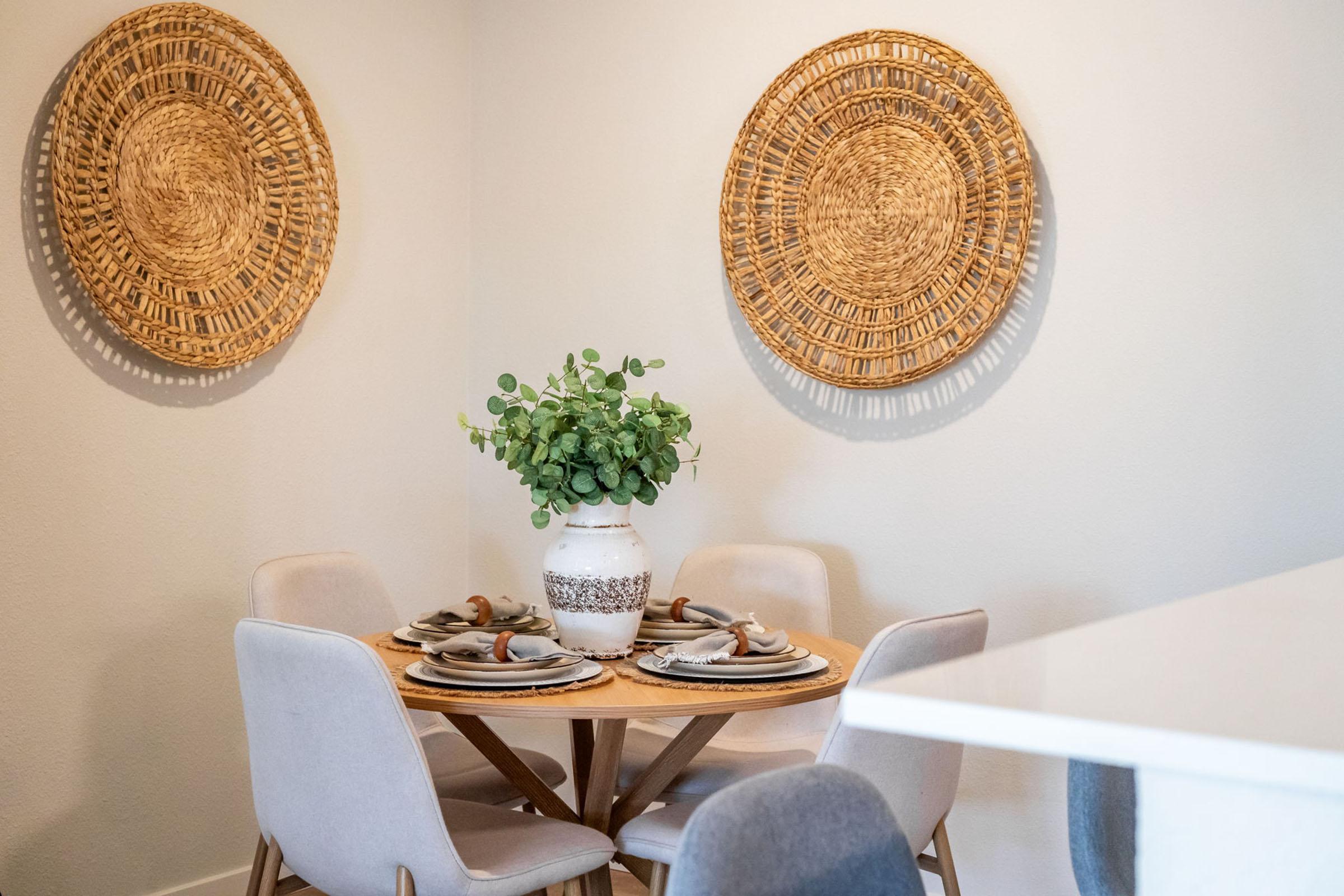 A cozy dining area featuring a round wooden table set with plates and cutlery, surrounded by four plush chairs. A decorative vase filled with greenery sits at the center, while two woven wall hangings add a warm touch to the beige wall. The overall ambiance is inviting and stylish.