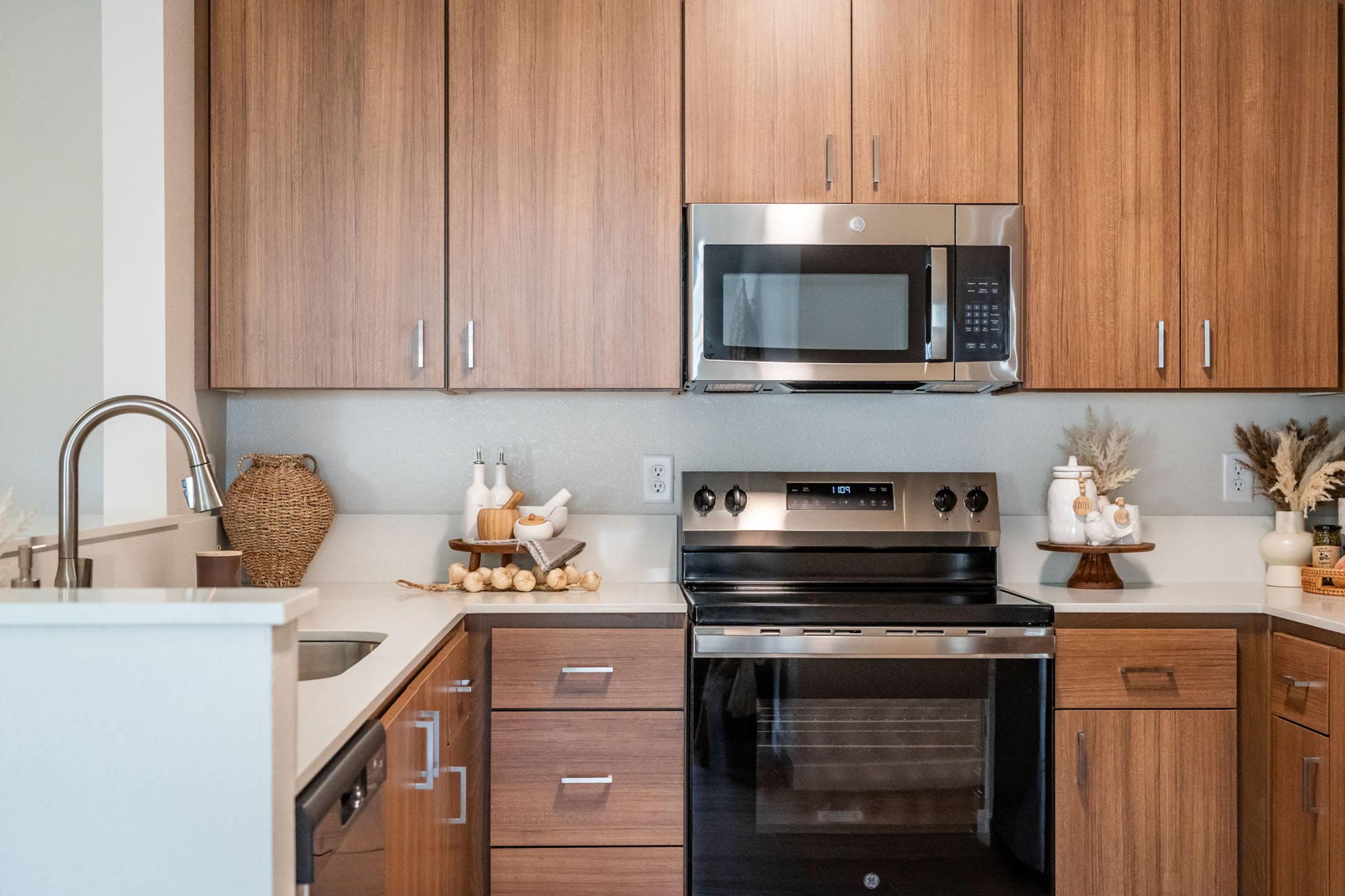 A modern kitchen featuring wooden cabinets, a stainless-steel microwave and oven, and a stylish sink. Decor elements include a woven basket, decorative bottles, and a bowl of wooden utensils, creating a warm and inviting atmosphere.