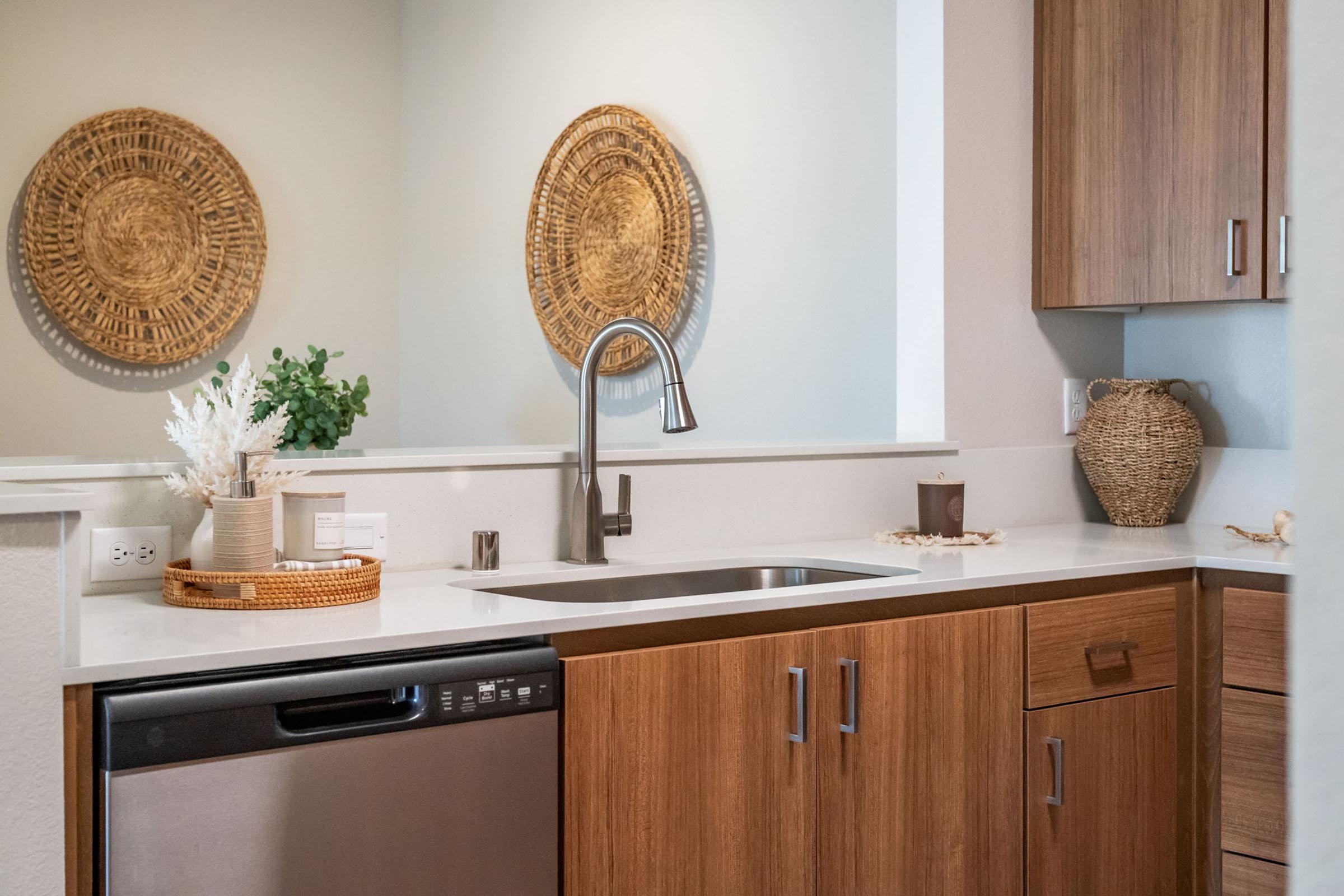 A modern kitchen featuring wooden cabinets, a stainless steel dishwasher, and a sleek sink. Decorated with woven baskets and circular wall hangings, the space has a light color scheme and a clean, minimalist aesthetic. A small plant and kitchen utensils are arranged on the counter.