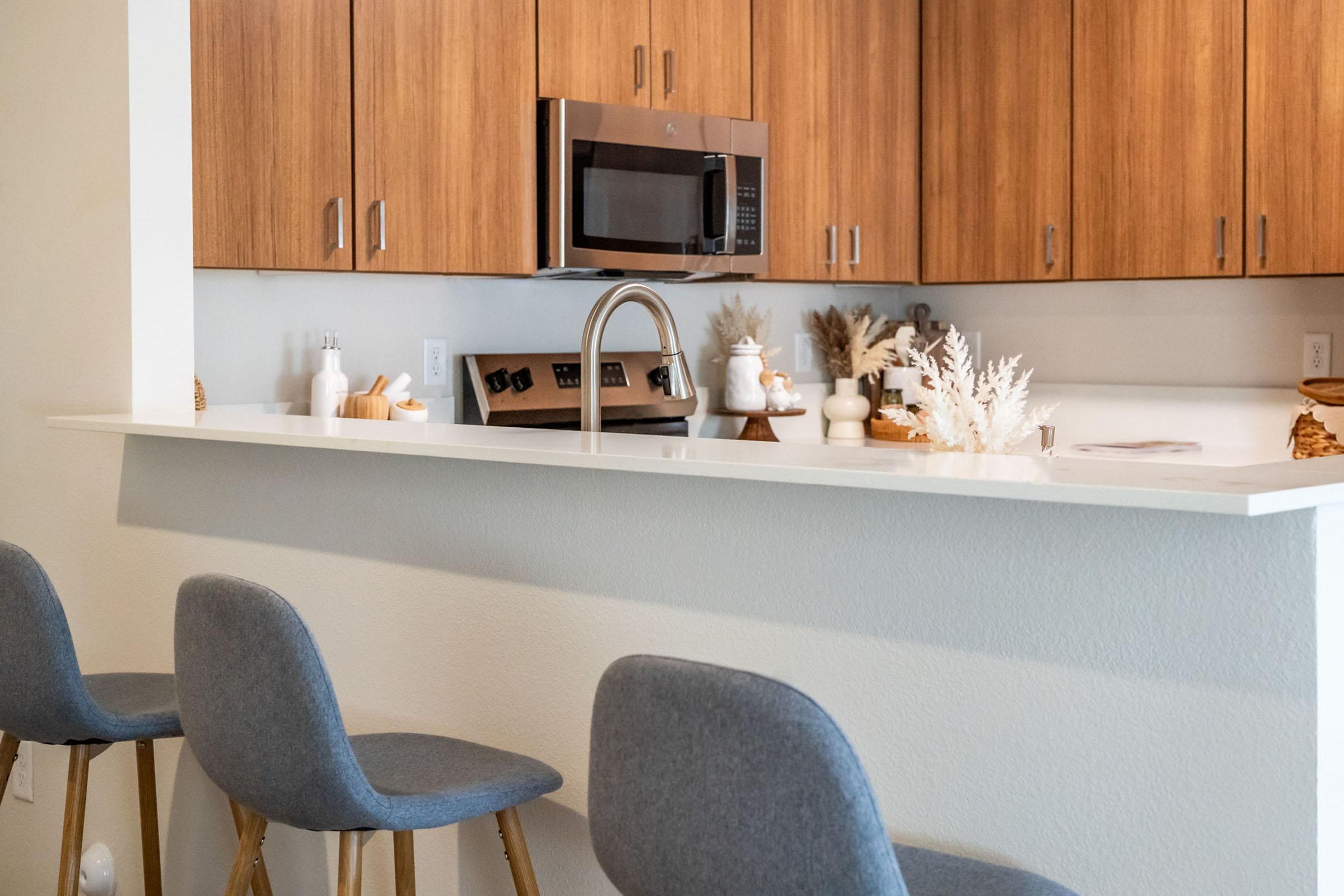 A modern kitchen with wooden cabinetry, a stainless steel microwave above a beige countertop. Bar stools with gray upholstery are positioned at the counter. Decorative items, including white vases and dried plants, are arranged on the counter for a stylish, inviting look.