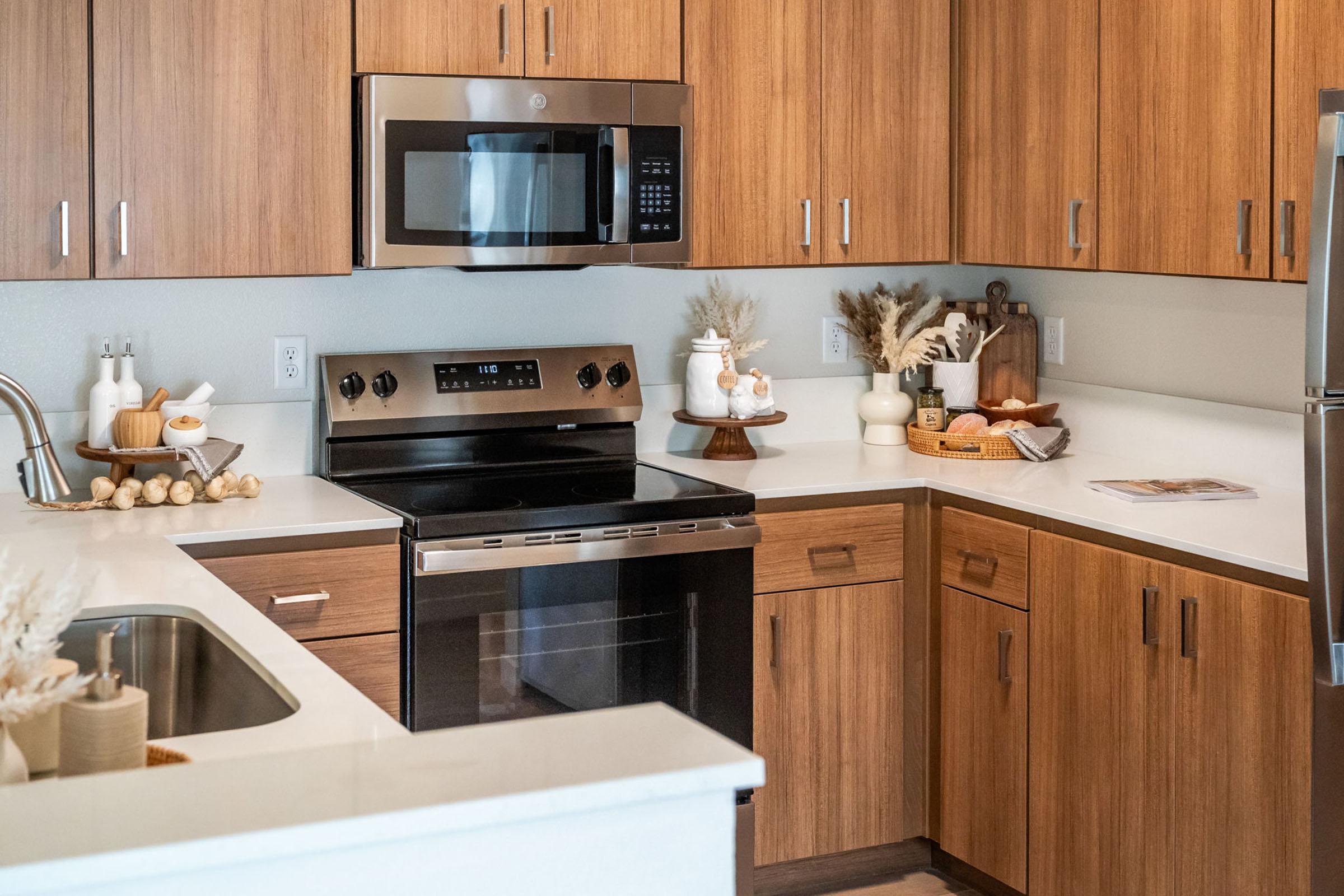 Modern kitchen featuring wooden cabinetry, a stainless steel microwave and stove, and a white countertop. Decorative items include a tray with dishes, a vase with dried flowers, and a small bowl. The overall design is warm and inviting with neutral tones and earthy accents.