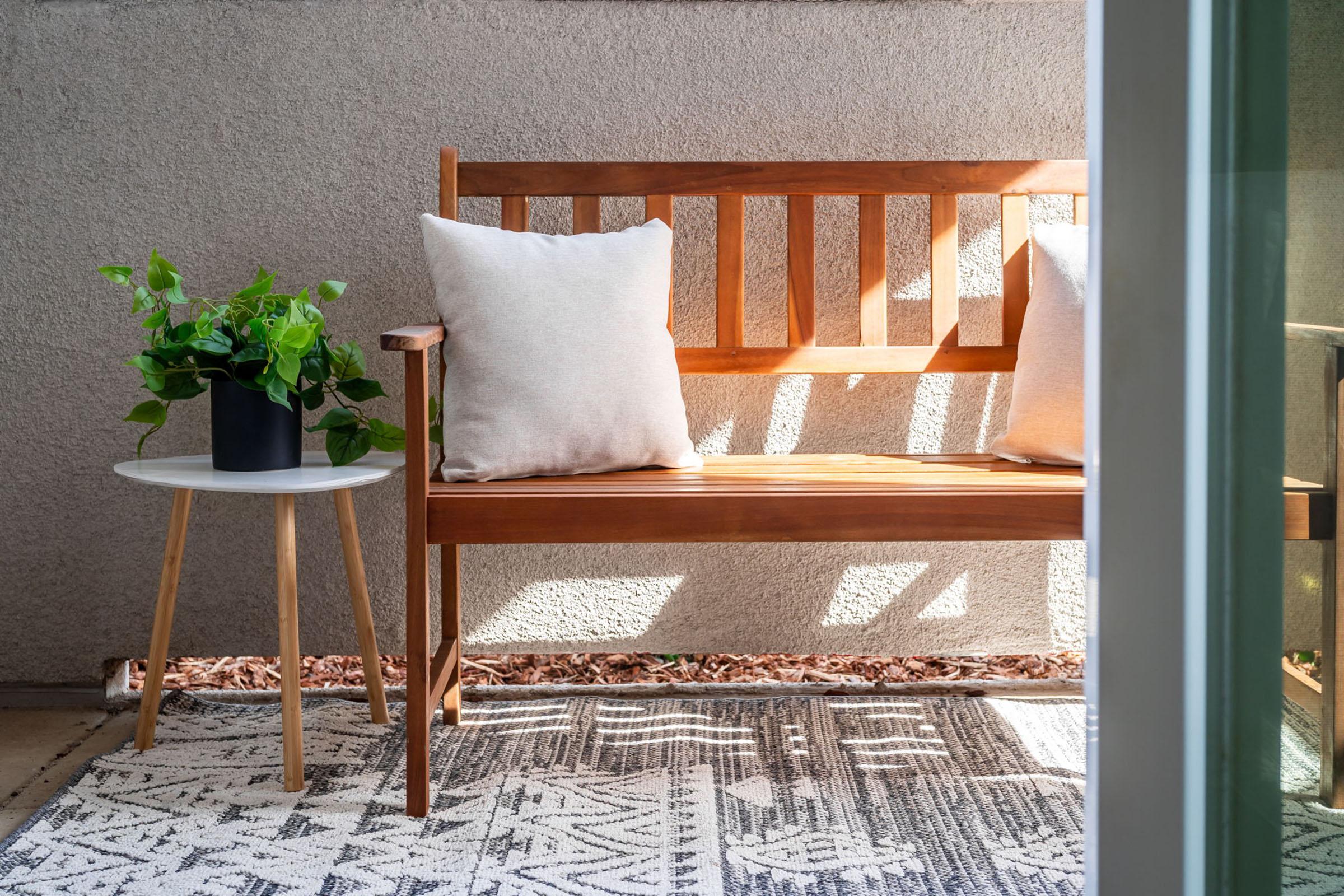 A wooden bench with two cushions sits next to a small round table on a patterned rug. A potted plant is on the table, and sunlight casts gentle shadows on the wall and floor, creating a serene outdoor seating area.