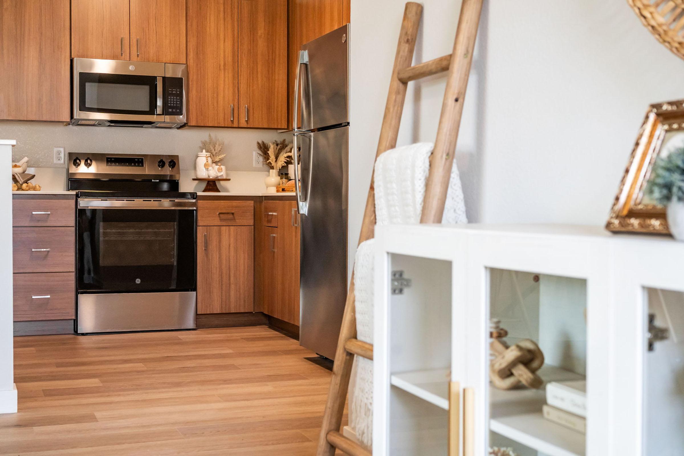 Modern kitchen featuring wooden cabinets, stainless steel appliances including a microwave and oven, and a refrigerator. A wooden ladder is leaning against the wall, and a white cabinet displays decorative items. The floor is light-colored wood, contributing to a warm, inviting atmosphere.
