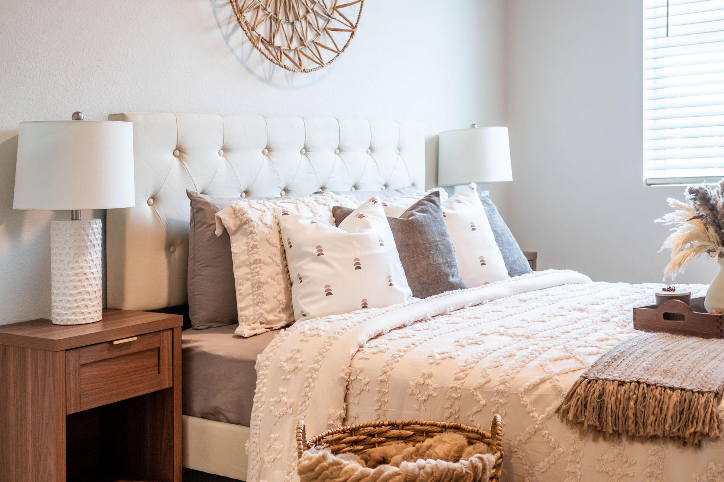 A cozy bedroom featuring a tufted beige headboard, decorative pillows on a neatly made bed with a cream-colored blanket. There are two bedside lamps on wooden nightstands, and a woven wall decor above the bed. Natural sunlight filters through a window, creating a warm, inviting atmosphere.