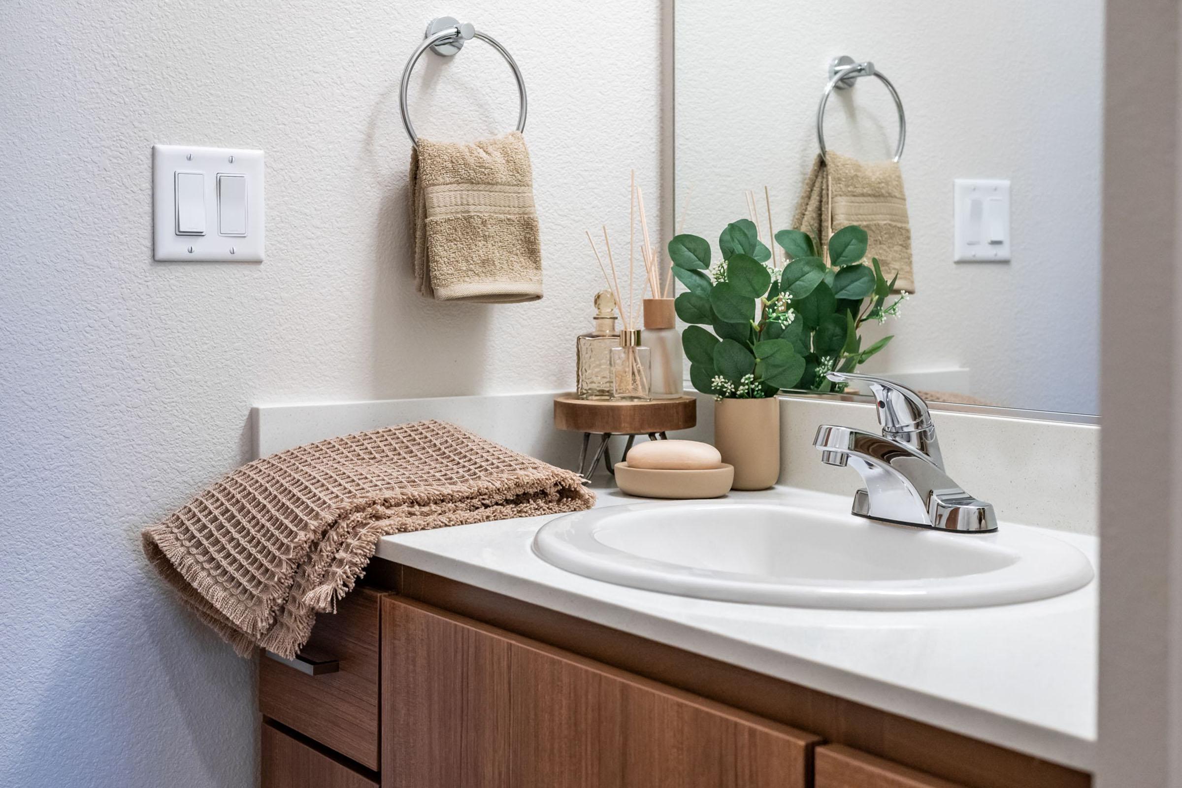 A clean and modern bathroom sink area featuring a chrome faucet, a small bowl, decorative plants, and a towel hanging on a ring. A woven towel is neatly draped nearby, and a round wooden tray holds various toiletries. The walls are painted a light color, enhancing the spacious feel.
