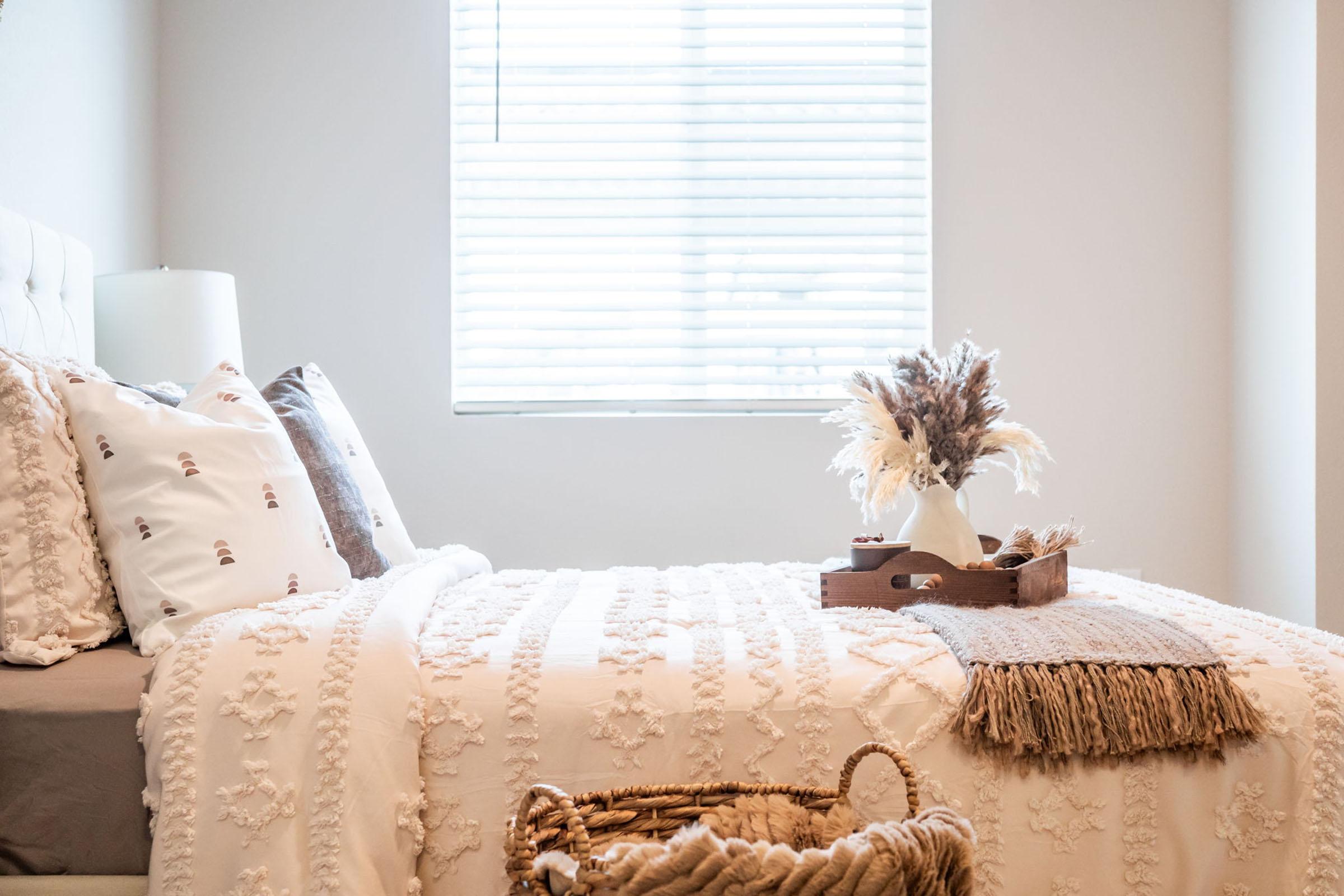 A cozy bedroom featuring a neatly made bed with decorative pillows and a textured blanket. A small tray with dried flowers and decorative items sits on the bed. A woven basket is on the floor, and soft natural light filters through a window with blinds, creating a warm and inviting atmosphere.