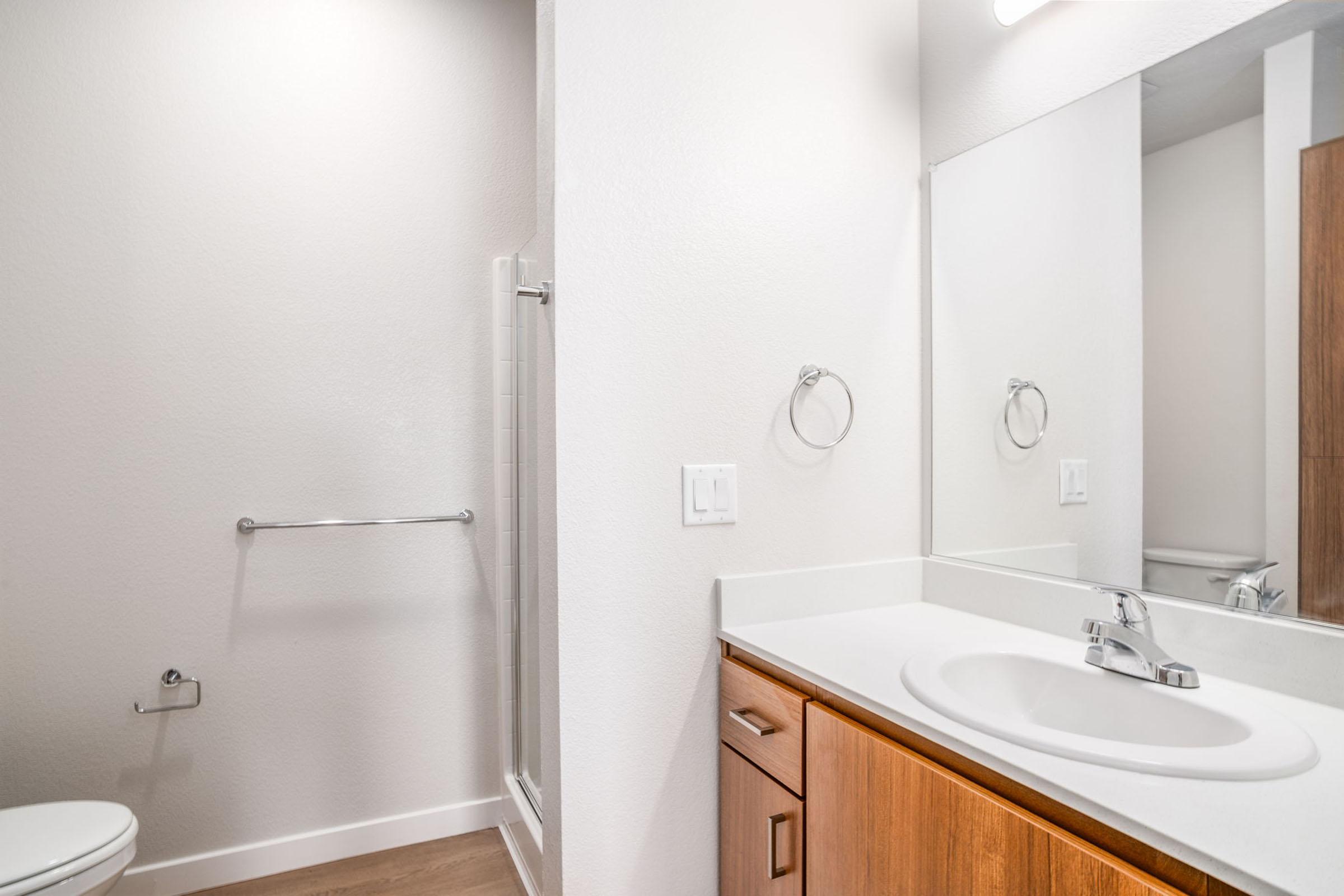Modern bathroom featuring a clean, light-colored design. The space includes a white countertop with a sink, a large wall-mounted mirror, wooden cabinetry, and a shower area. A towel bar and toilet can also be seen, with neutral tones creating a bright, airy atmosphere.