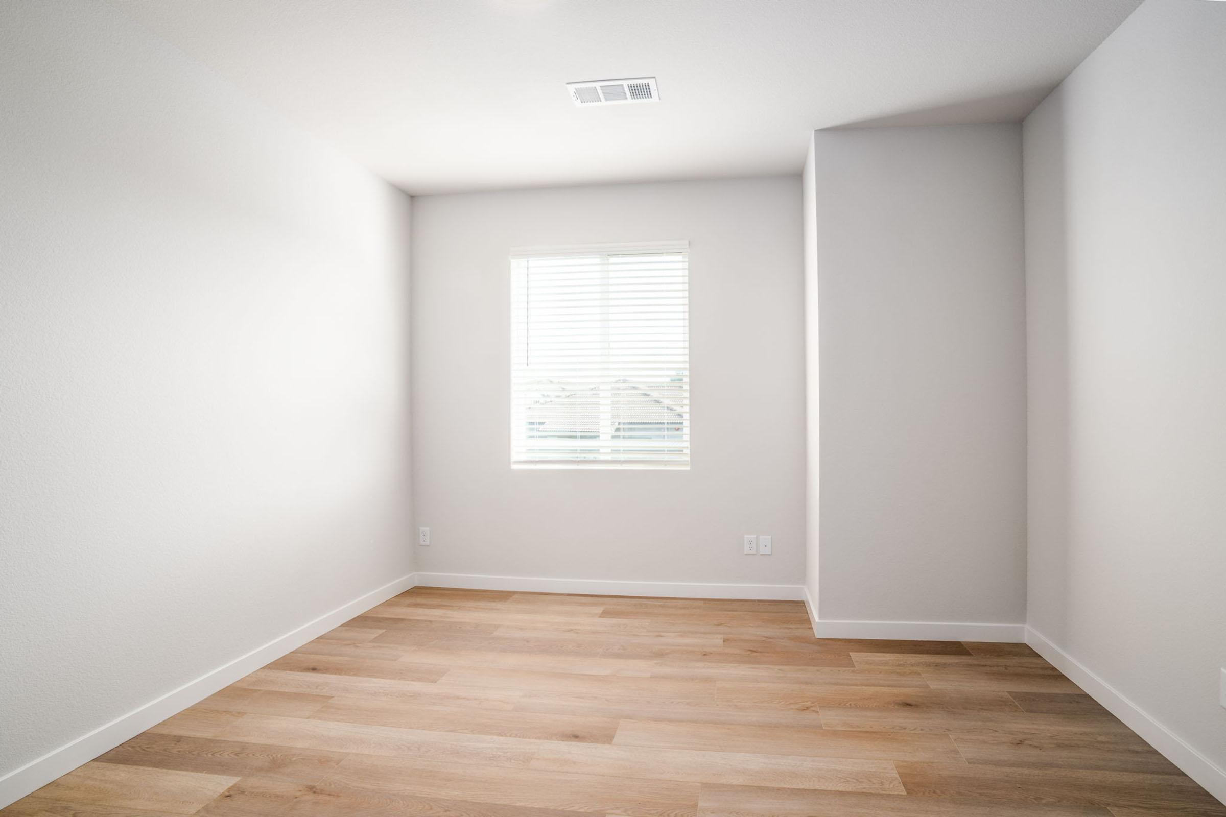 A bright, empty room with light-colored walls and wooden flooring. A single window features white blinds, allowing natural light to enter. The space is minimalistic, designed for various uses, with no furniture present.