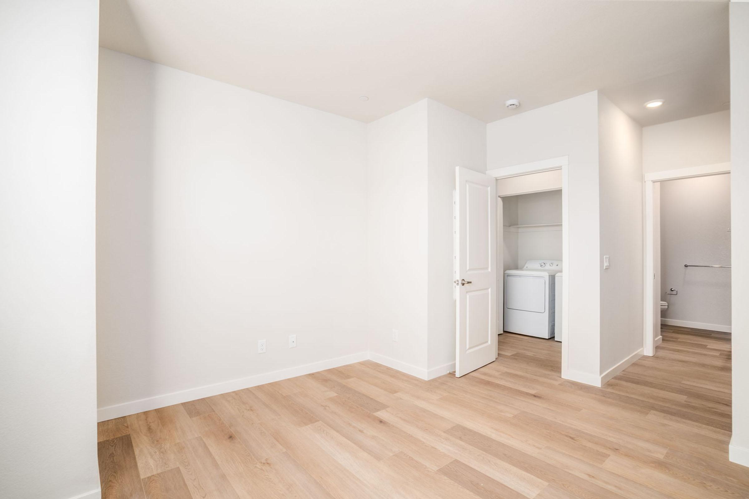 A spacious, empty room with light-colored walls and wooden flooring. A door on the left leads to a small laundry area with a washer, while another doorway on the right leads to a bathroom. The room has natural light and a clean, minimalist design.