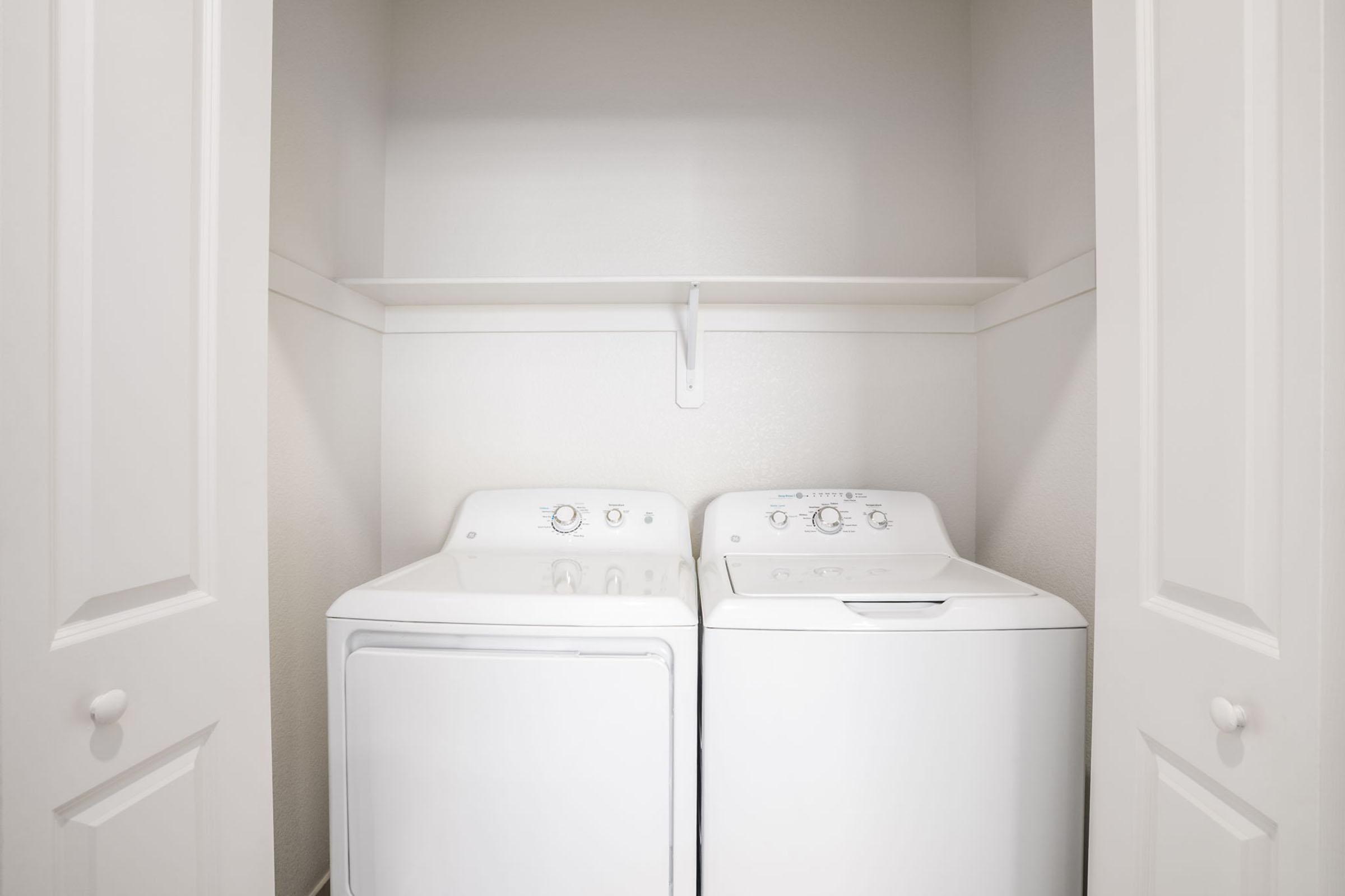 A clean and tidy laundry room featuring a side-by-side washer and dryer, enclosed within white closet doors. The walls are painted light, and a shelf is mounted above the appliances for added storage.