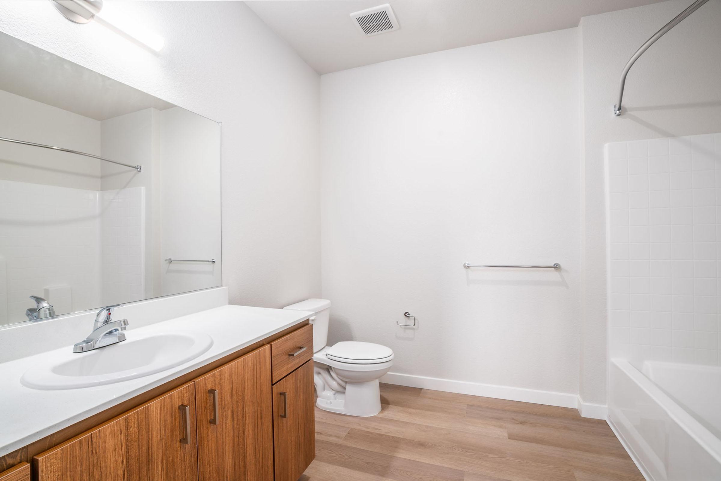 Modern bathroom featuring a large mirror, wooden vanity with a sink, a toilet, and a bathtub with a shower. The walls are painted white, and the floor has light wooden planks. Natural light comes from a ceiling fixture, creating a bright and airy atmosphere.