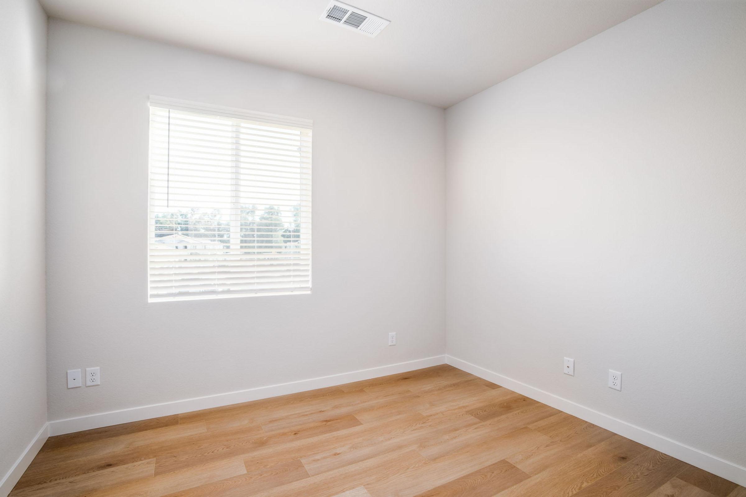 Empty room with light-colored walls and wooden flooring. A window with blinds lets in natural light, illuminating the space. The room has minimal furnishings, creating a clean and open atmosphere.