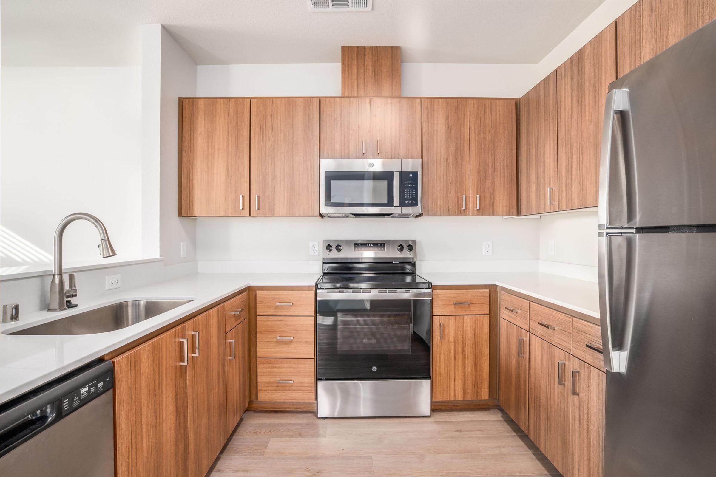 Modern kitchen featuring wooden cabinetry, stainless steel appliances including a microwave, stove, and refrigerator, and a double basin sink. The countertops are white, and the flooring is light wood. Natural light illuminates the space from a window, creating a bright and inviting atmosphere.