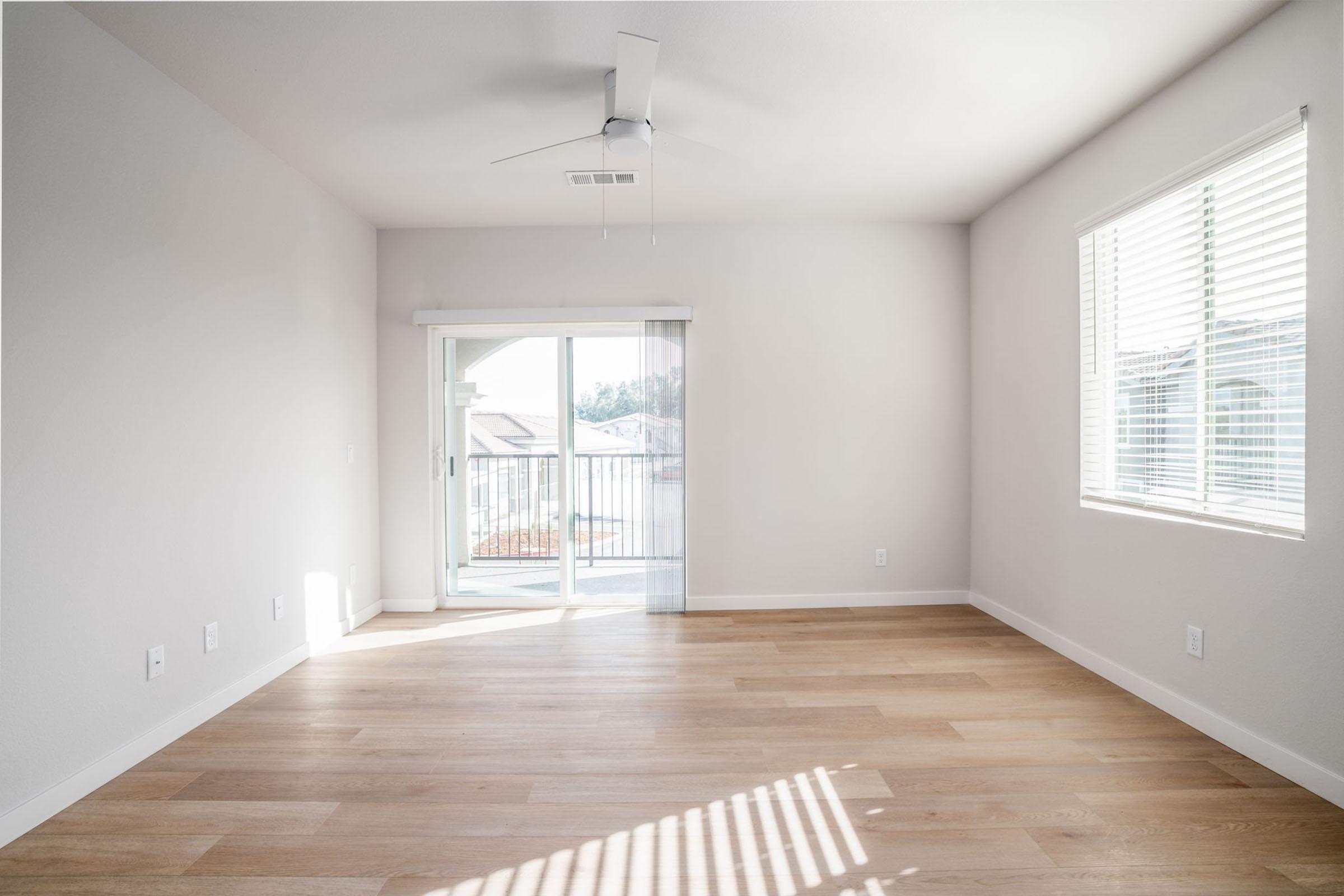 A spacious, empty room with light gray walls and new wooden flooring. A ceiling fan is mounted on the flat ceiling, and a sliding glass door with blinds leads to a balcony. Natural light streams in through the window on the right. The room has a minimalist, modern design.