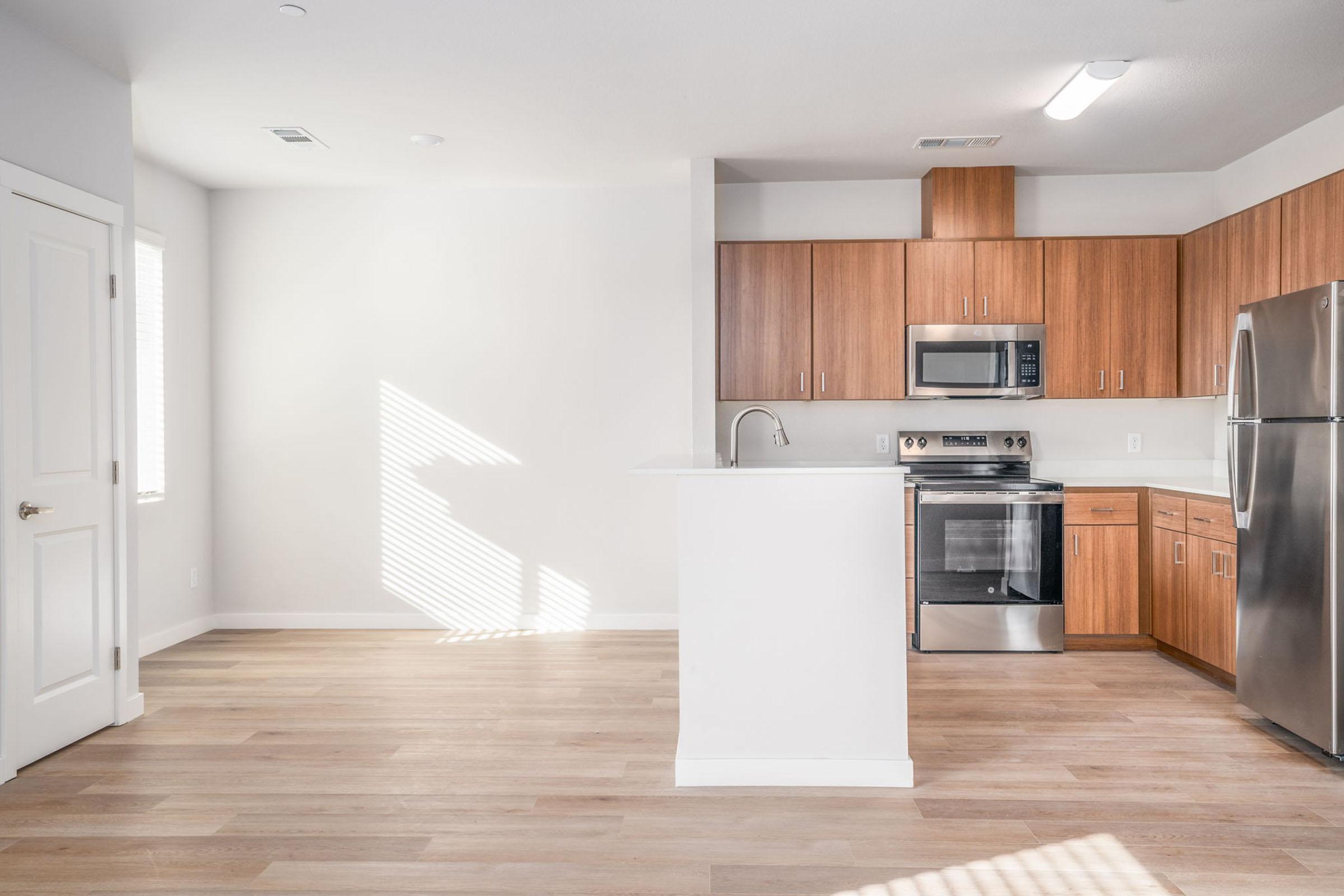 Modern kitchen with wooden cabinetry, stainless steel appliances, and a white countertop. The space features large windows allowing natural light to brighten the room, with light wooden flooring and minimalistic design elements creating an open and airy feel.