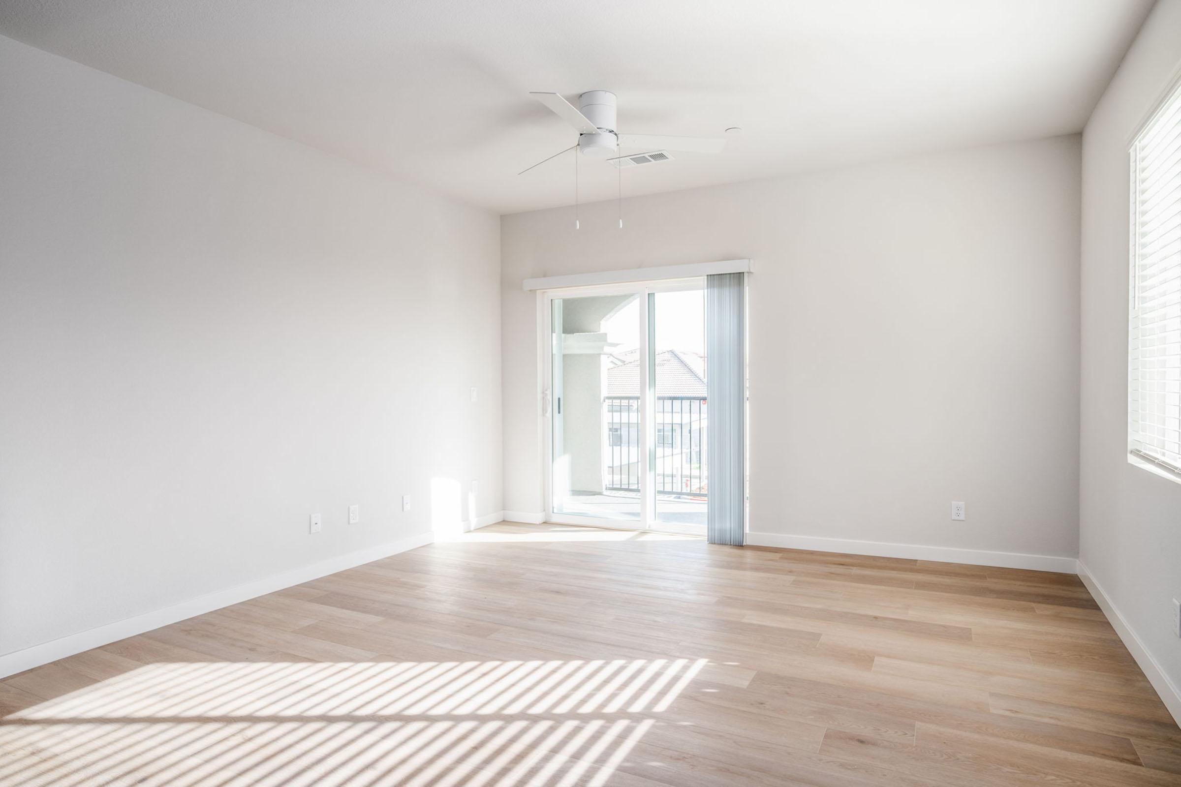 Bright, empty living room with light-colored walls and hardwood flooring. A ceiling fan is mounted on the ceiling, and large sliding glass doors lead to a balcony, allowing natural light to flood the space. Sunlight creates striped patterns on the floor from the window blinds.
