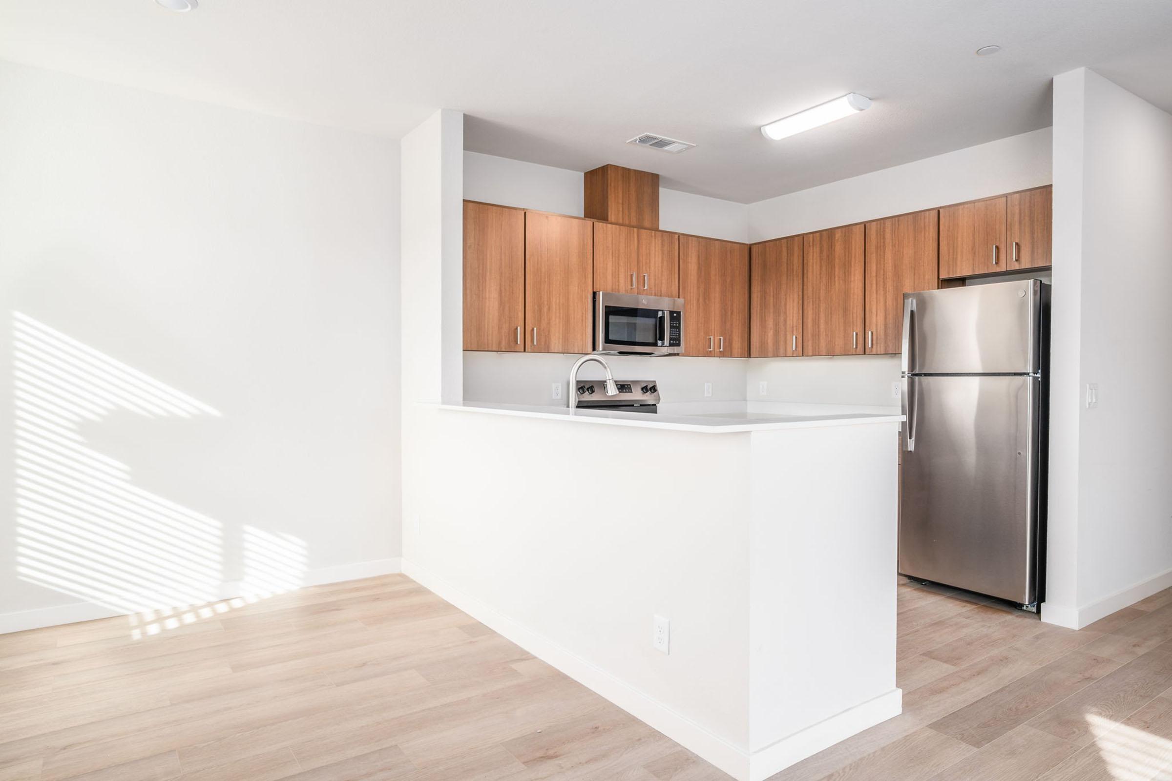 Bright and modern kitchen featuring wooden cabinetry, stainless steel refrigerator, and microwave. A large kitchen island with a sink offers a spacious layout, complemented by light-colored walls and warm wood flooring. Natural light streams through a window, creating an inviting atmosphere.
