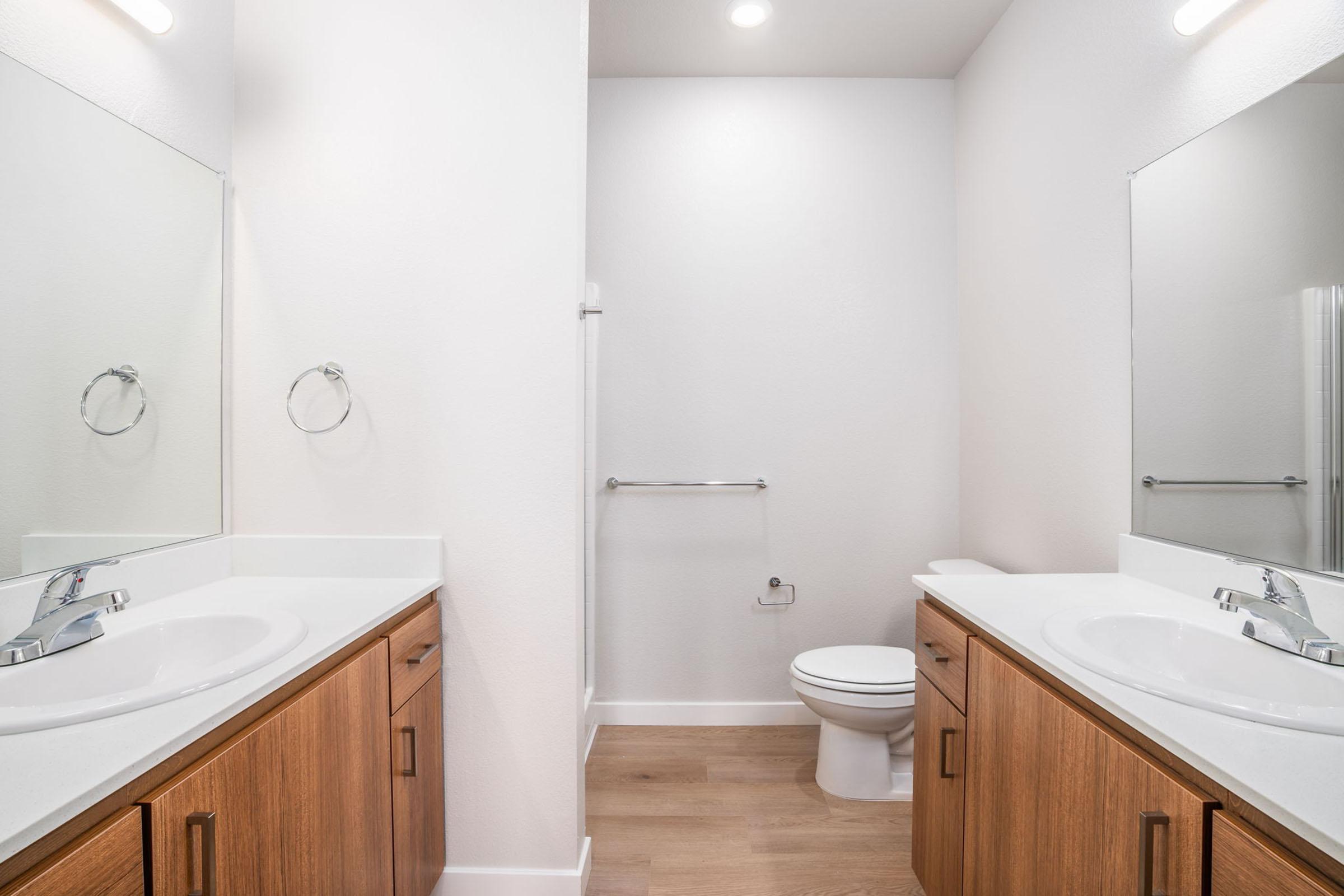 A modern bathroom featuring two sinks with chrome faucets, wooden cabinetry, a toilet, and a walk-in shower. The walls are painted in a light color, and there are two large mirrors above the sinks. The floor is finished in light wood. The overall design is clean and contemporary.
