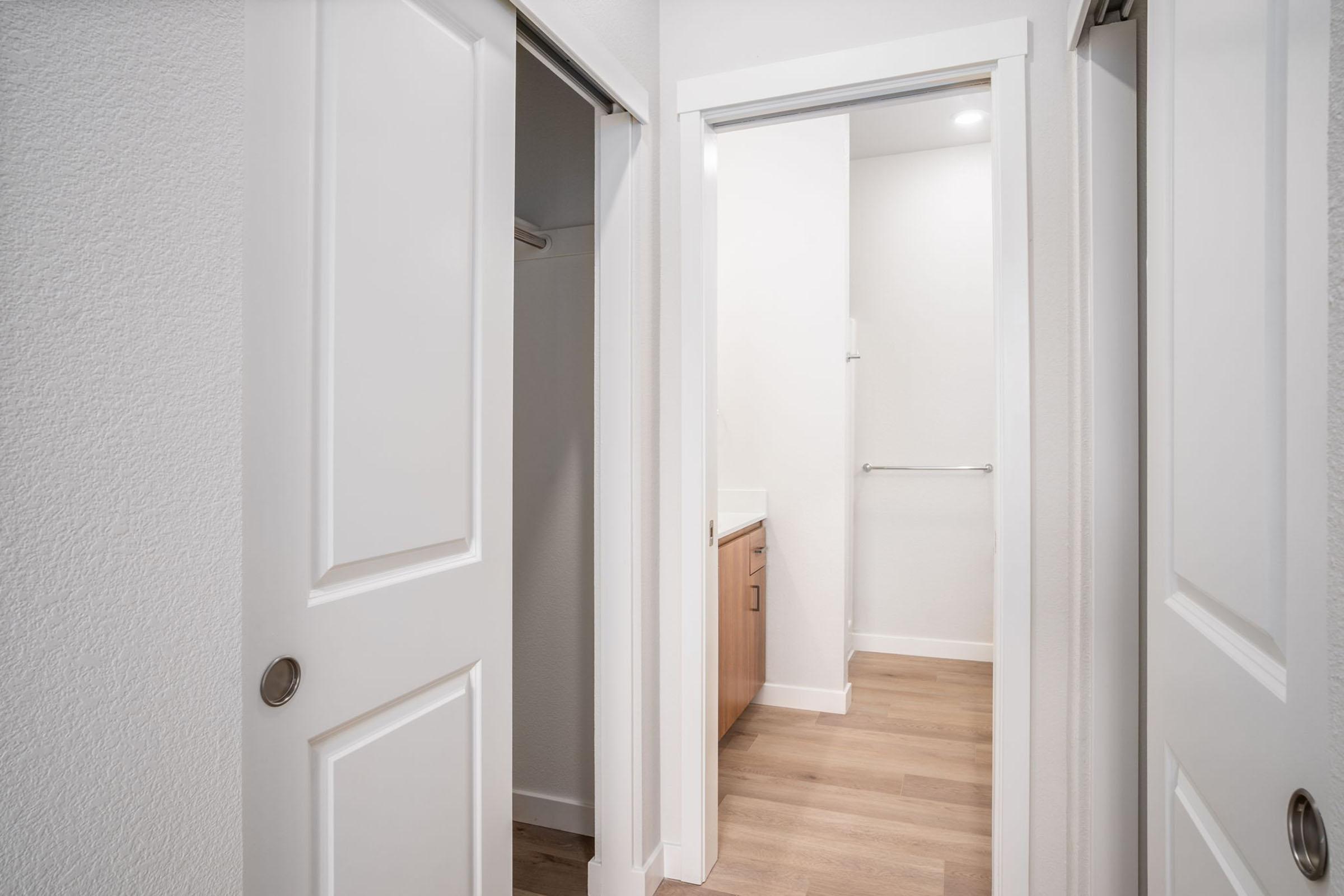 View of a small hallway featuring two doors. One door leads to a closet, while the other opens to a bathroom area with light-colored cabinetry. The floor is wooden, and the walls are painted white, creating a bright and modern atmosphere.