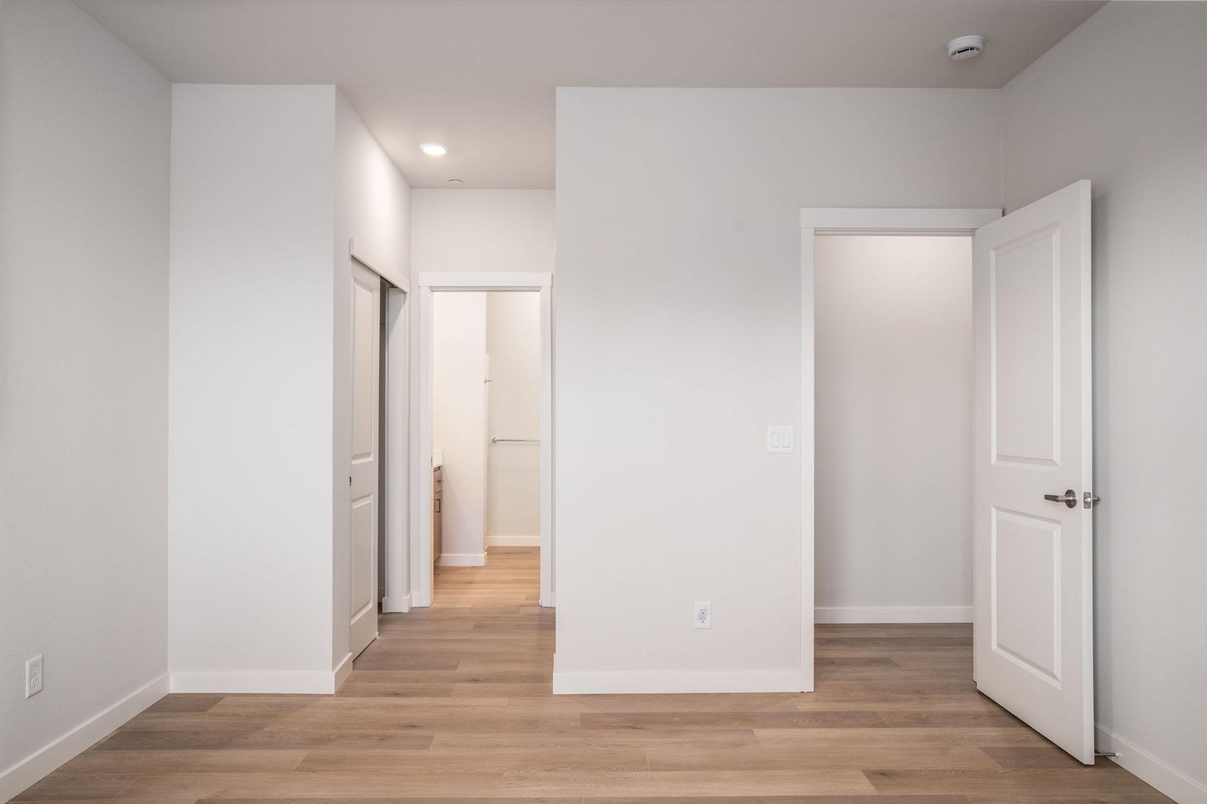 A light, modern hallway with white walls and light wood flooring. There are three doorways: one on the left leading to a room, a narrow one straight ahead, and another door on the right. The space has a bright, open feel with natural light.