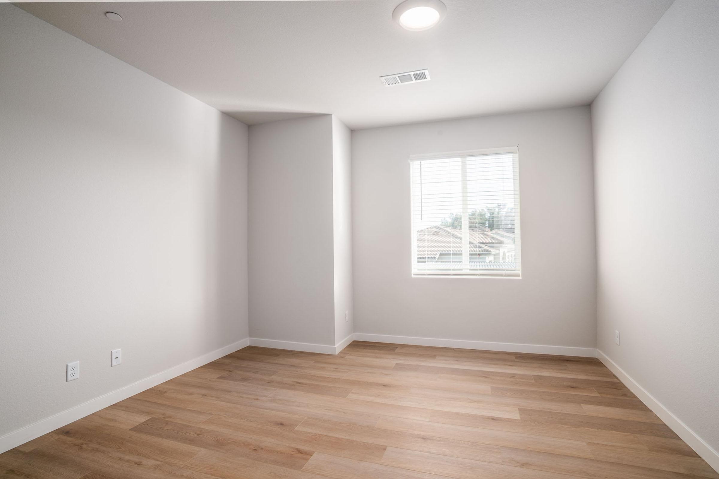 A modern, empty room featuring light gray walls, hardwood flooring, and a single window with horizontal blinds allowing natural light. The space has a corner nook and a ceiling light fixture, creating a bright and airy atmosphere, ideal for various uses such as an office or bedroom.