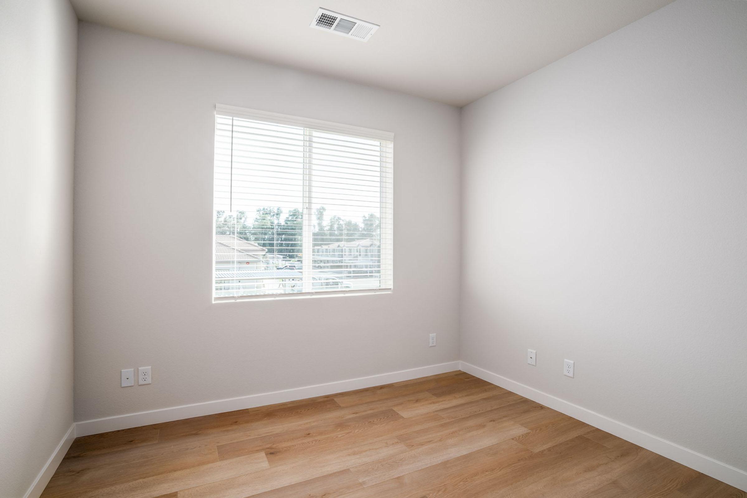 A bright, empty room with light-colored walls and wooden flooring. A large window with white blinds allows natural light to enter, illuminating the space. The room features a simple and modern design, with no furniture present, creating a minimalistic look.