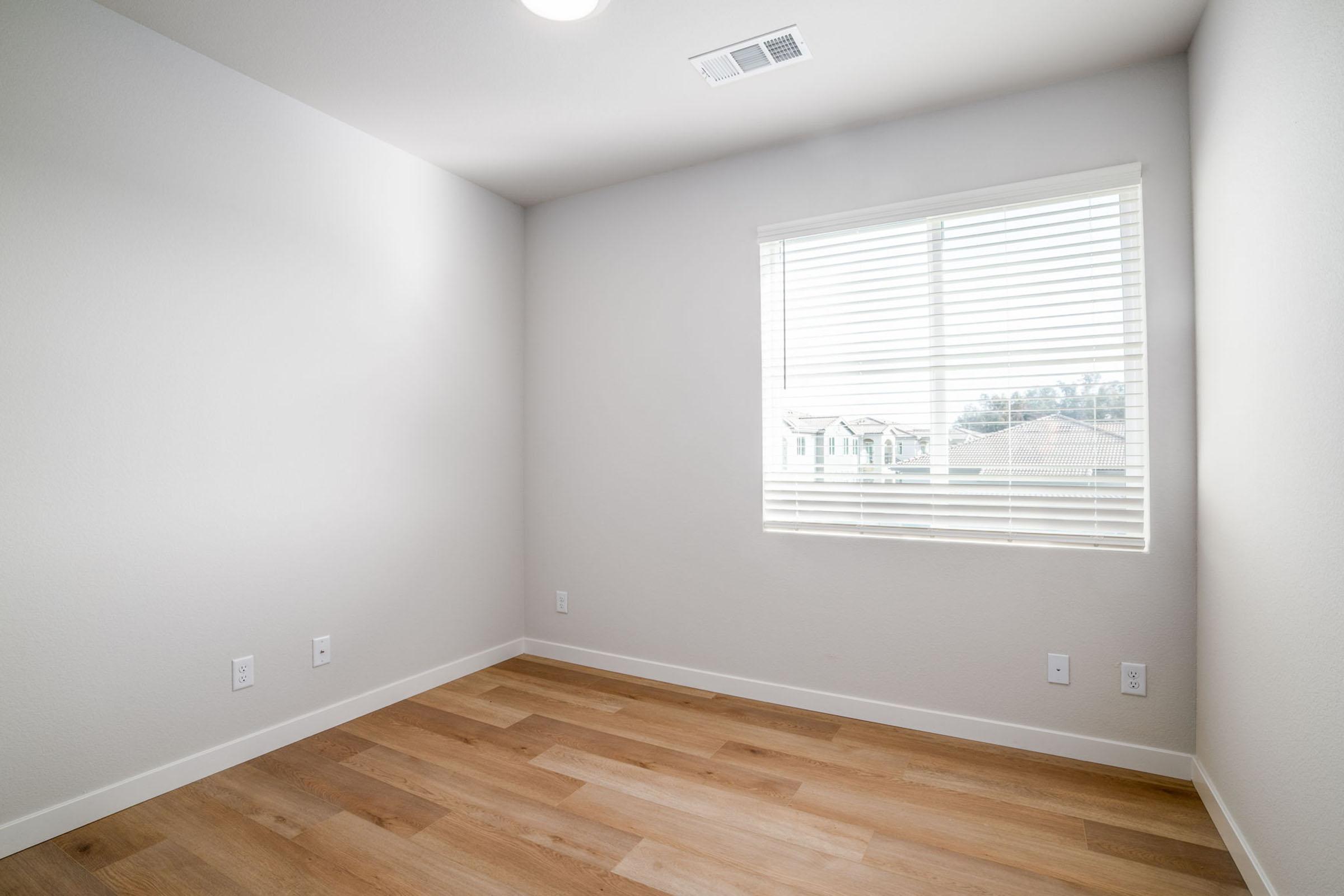 A light and airy empty room featuring neutral gray walls and wooden flooring. There's a large window with white blinds that allows natural light to enter, offering a view of the outdoors. The space is clean and uncluttered, suitable for various uses such as a bedroom or office.