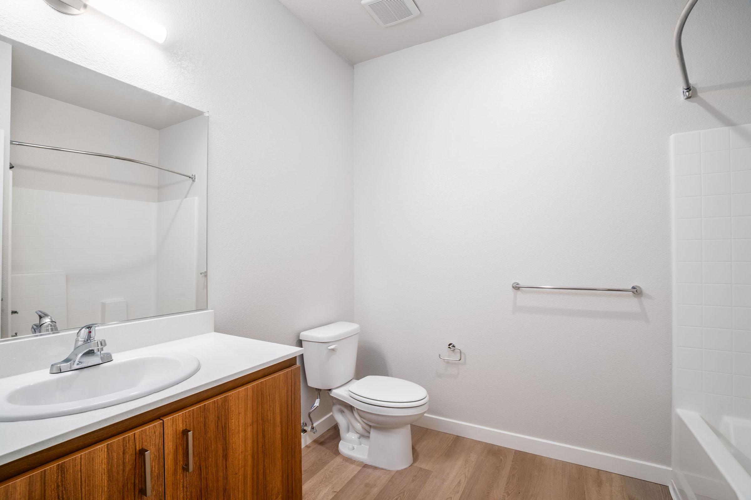 A modern, clean bathroom featuring a white sink with a mirror above, a white toilet, and a bathtub with a shower. The walls are painted a light color, and the floor has light-colored wood or laminate. There is a handrail next to the bathtub and a small cabinet beneath the sink.