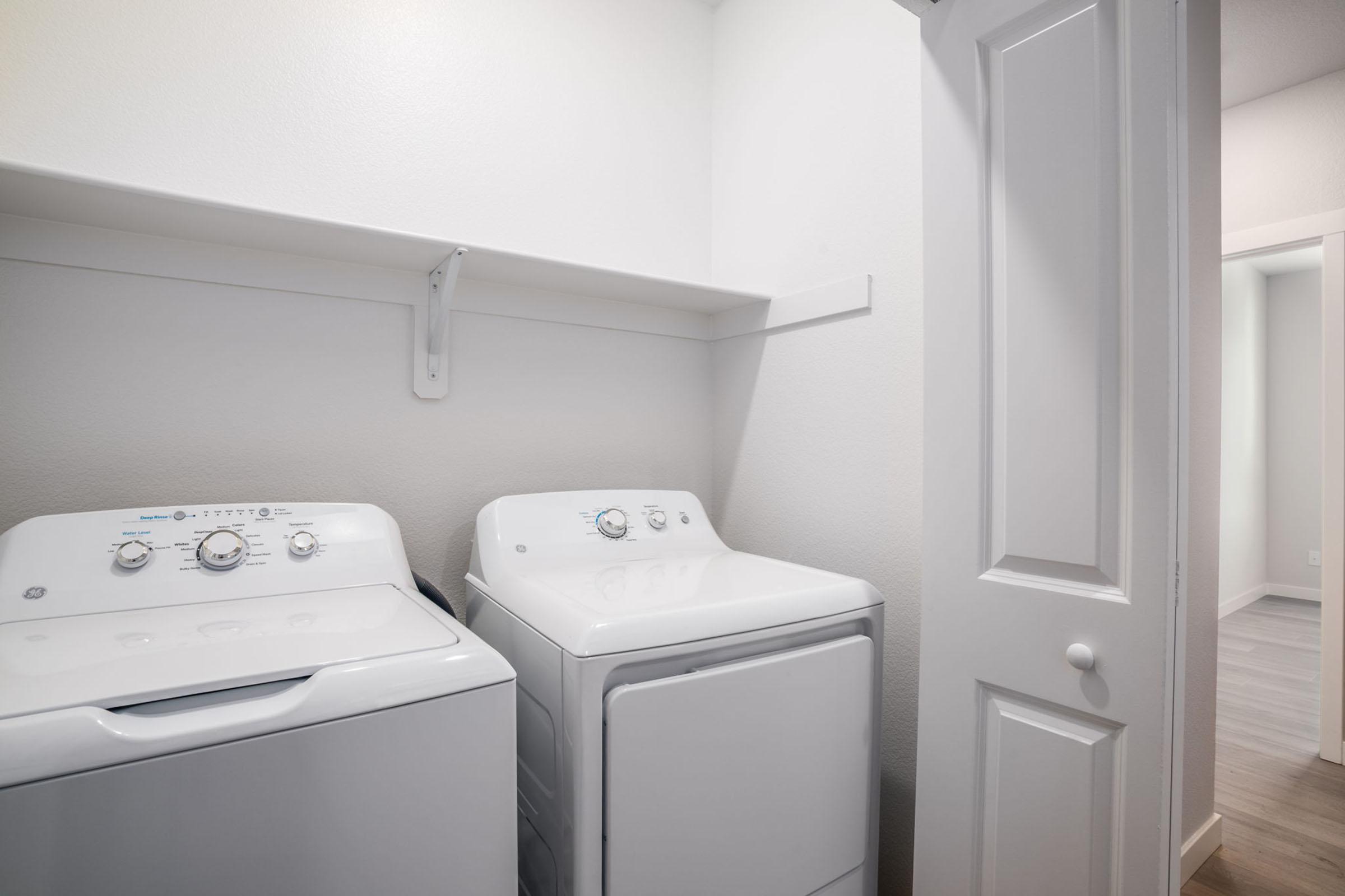 A clean laundry room featuring a white washing machine and dryer side by side, positioned against a light-colored wall. A small shelf is mounted above the appliances, and a door is visible on the right side of the image, leading to another room. The flooring is light-colored wood.