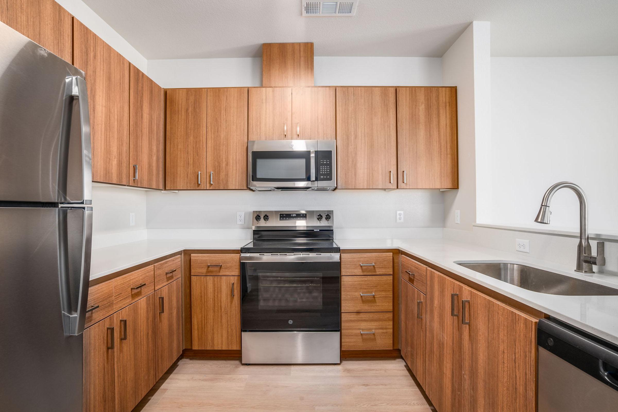 Modern kitchen featuring wooden cabinets, stainless steel appliances, including a refrigerator and oven, with a microwave above. The countertops are light-colored, and there is a double sink. The space is bright and contemporary, with an open layout and minimalistic design.