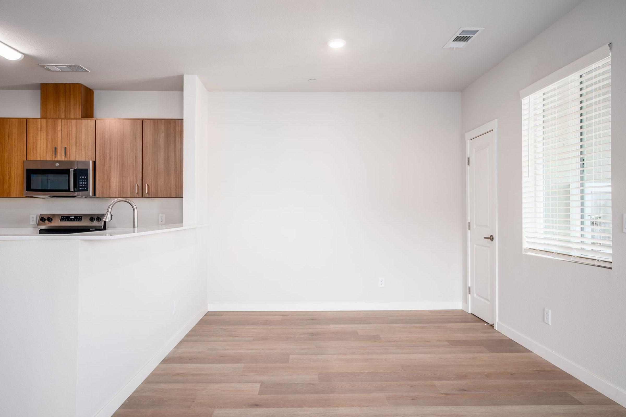 A modern, unfurnished kitchen and living area featuring light wood cabinetry, a stainless steel microwave, and an open space with hardwood flooring. Natural light streams in through a window, and a white door leads to another room, showcasing a clean and minimalist design.