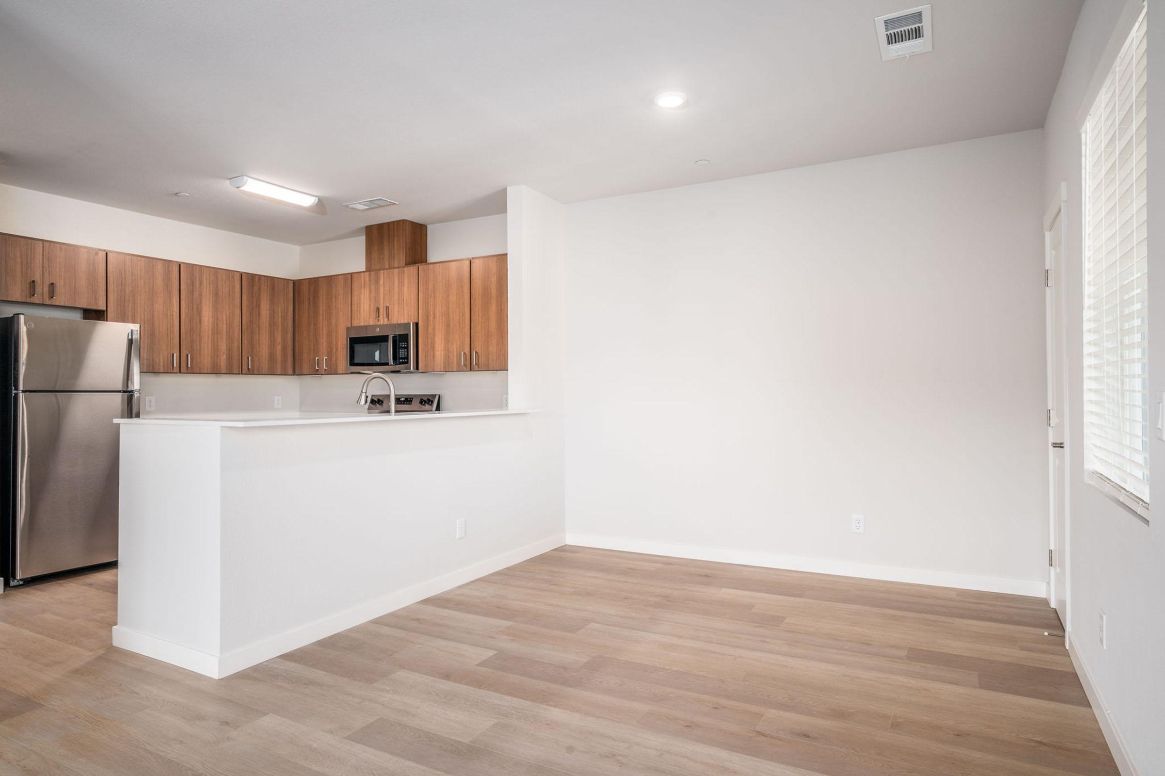 A modern kitchen space featuring light wood cabinetry, stainless steel appliances, and an open layout. The hardwood floor extends into a spacious living area with light walls and a window. The design is minimalistic and bright, emphasizing a contemporary aesthetic.