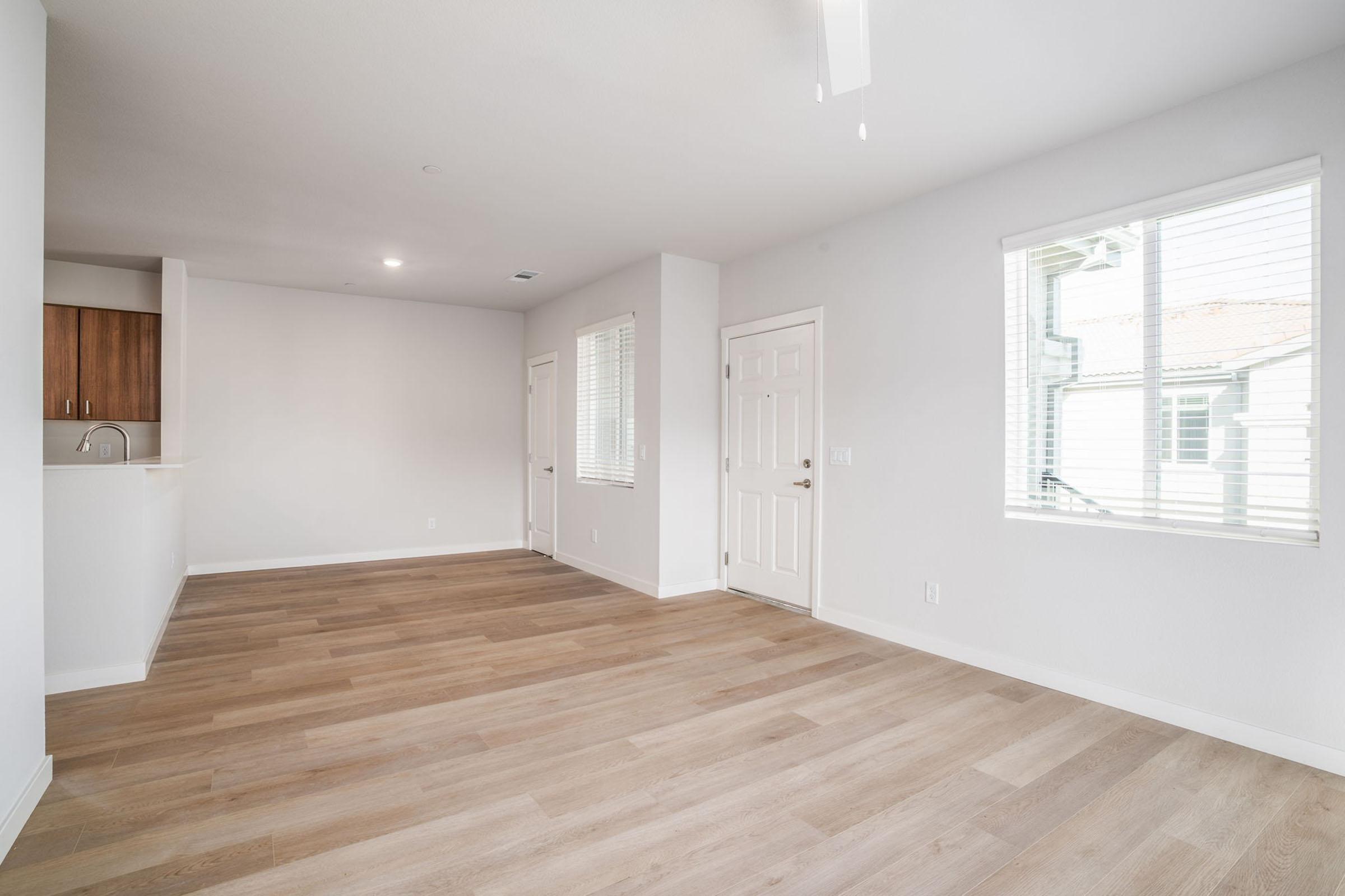 Empty living space with light-colored walls and wood-like flooring, featuring a window with blinds, a ceiling fan, and a door leading outside. The adjacent area shows a partial view of a kitchen with wooden cabinets. The room is bright and airy, providing a sense of openness.