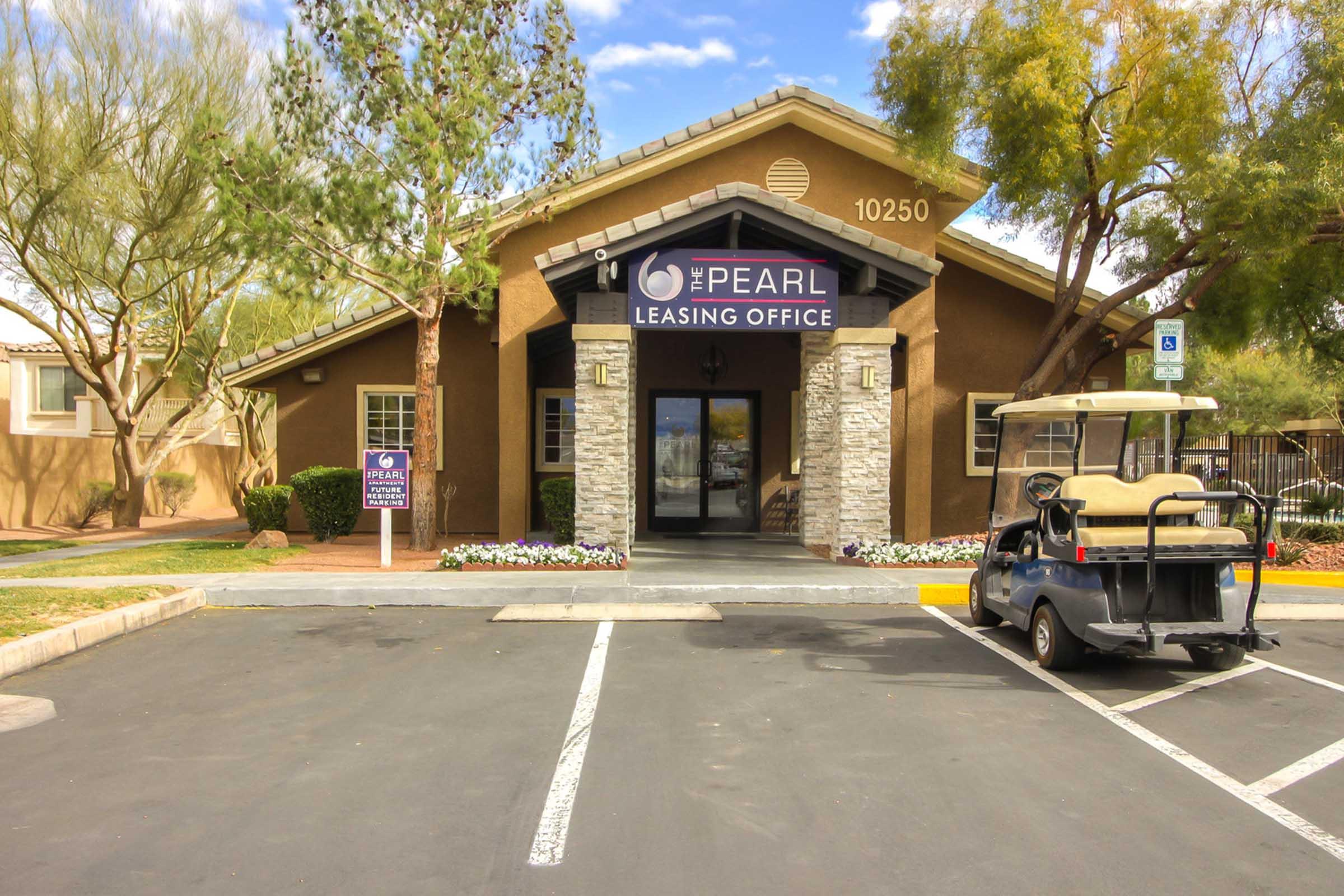 Leasing office exterior for The Pearl, featuring a stone and wood entrance, sign displaying "The Pearl Leasing Office," vibrant landscaping, a golf cart parked nearby, and a clear sky in the background.