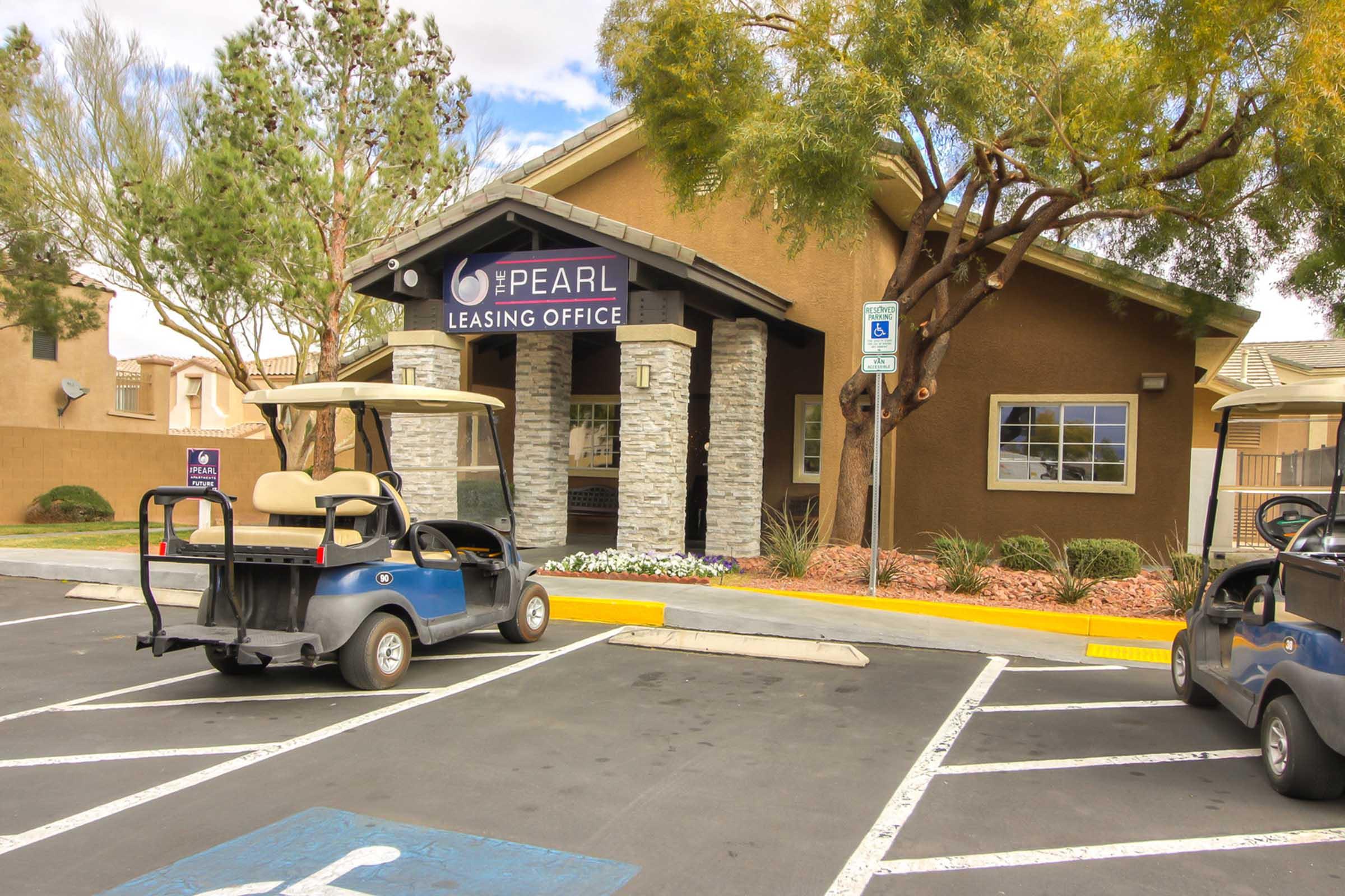 Leasing office of The Pearl with a sign, surrounded by landscaped areas. Two golf carts parked in front, one in a disabled parking spot. The building features a stone façade and is situated in a residential neighborhood. Trees and shrubs add to the exterior aesthetics.