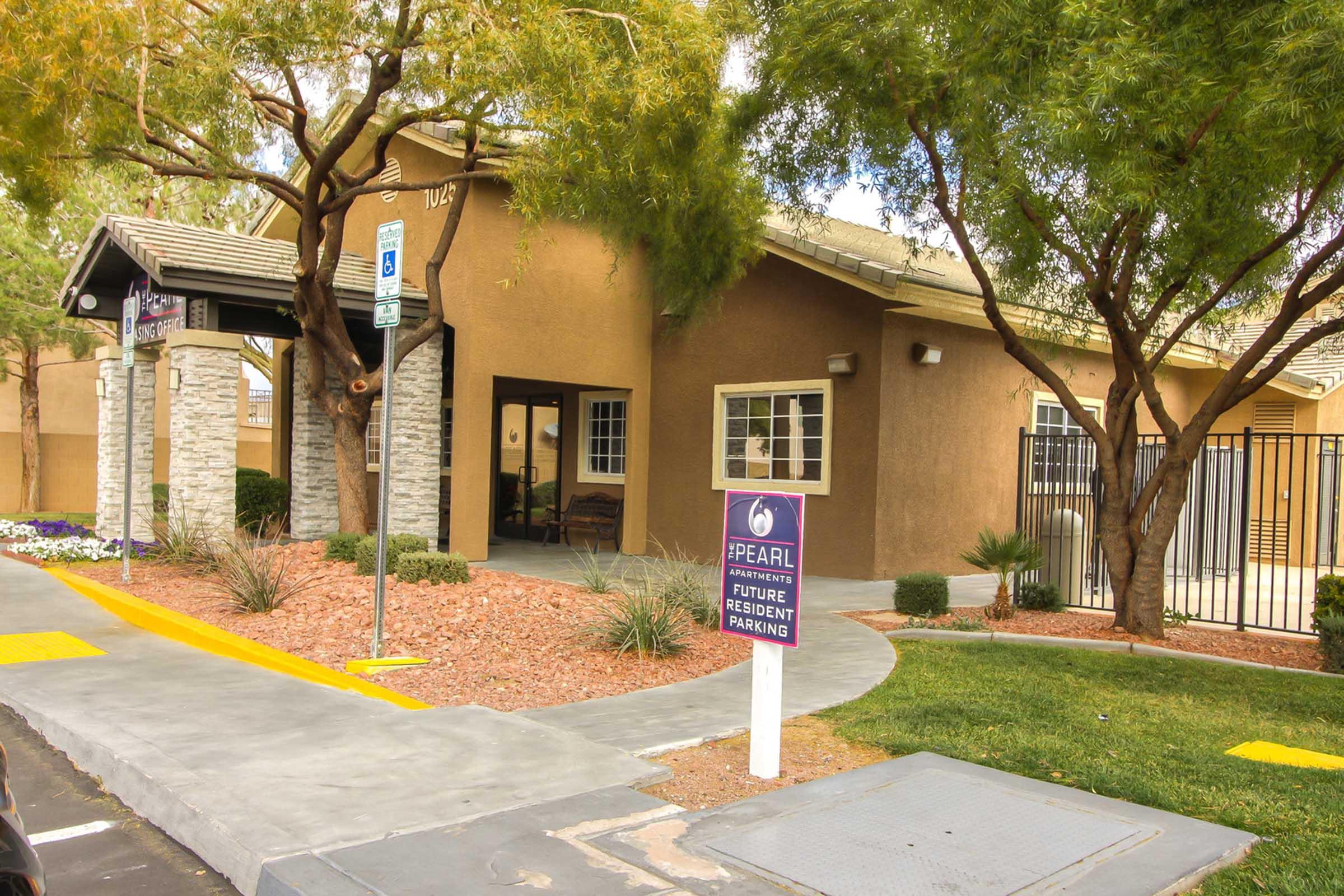 A view of an apartment complex entrance with a stone-accented façade and landscaped surroundings. The pathway leads to a door, and a sign indicates "Future Resident Parking." The area features greenery, flowers, and a gated section.