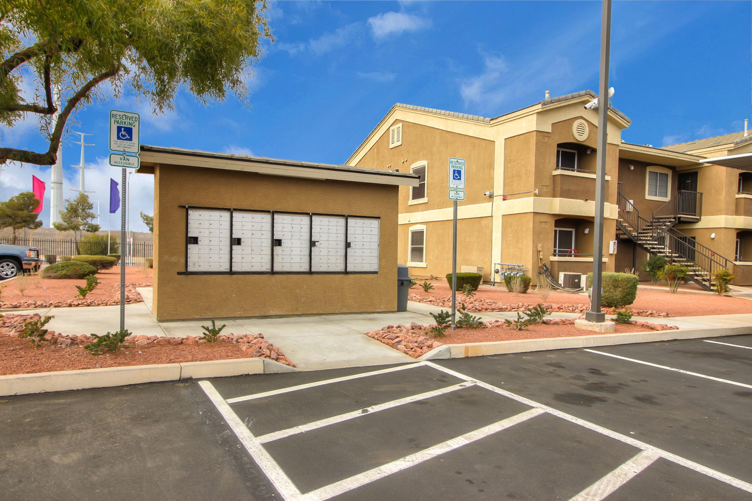 A beige outdoor mail station with multiple mailbox slots in front of a tan apartment building. Surrounding the area are landscaped features, including small shrubs and decorative rocks, with a clear blue sky and a few parking spaces visible in the foreground.