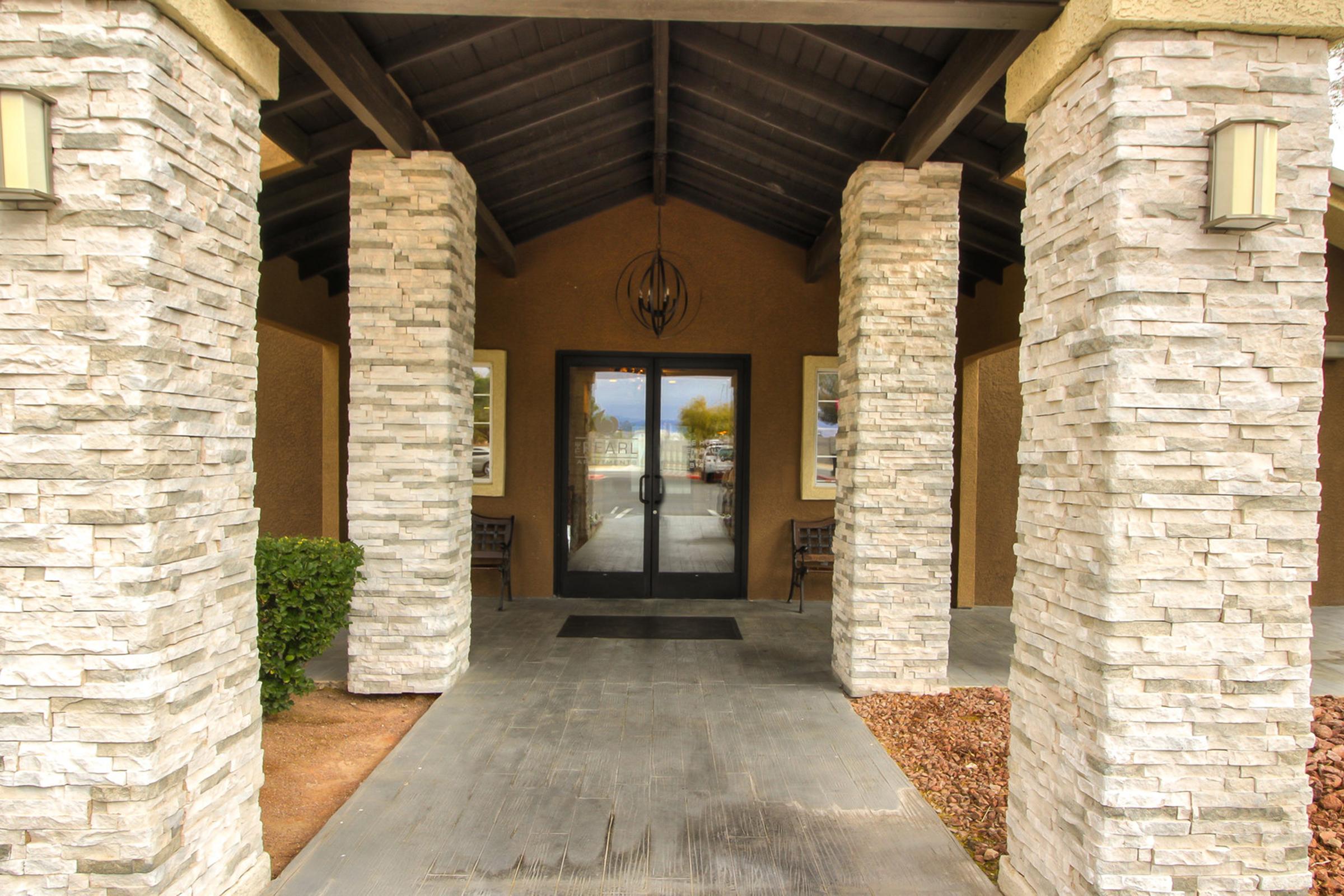 Entrance of a building featuring large stone columns supporting a dark wooden overhang. Double glass doors provide access, framed by stone pillars. There are benches on either side and decorative lighting fixtures mounted on the walls. Surrounding landscape includes gravel and shrubbery.