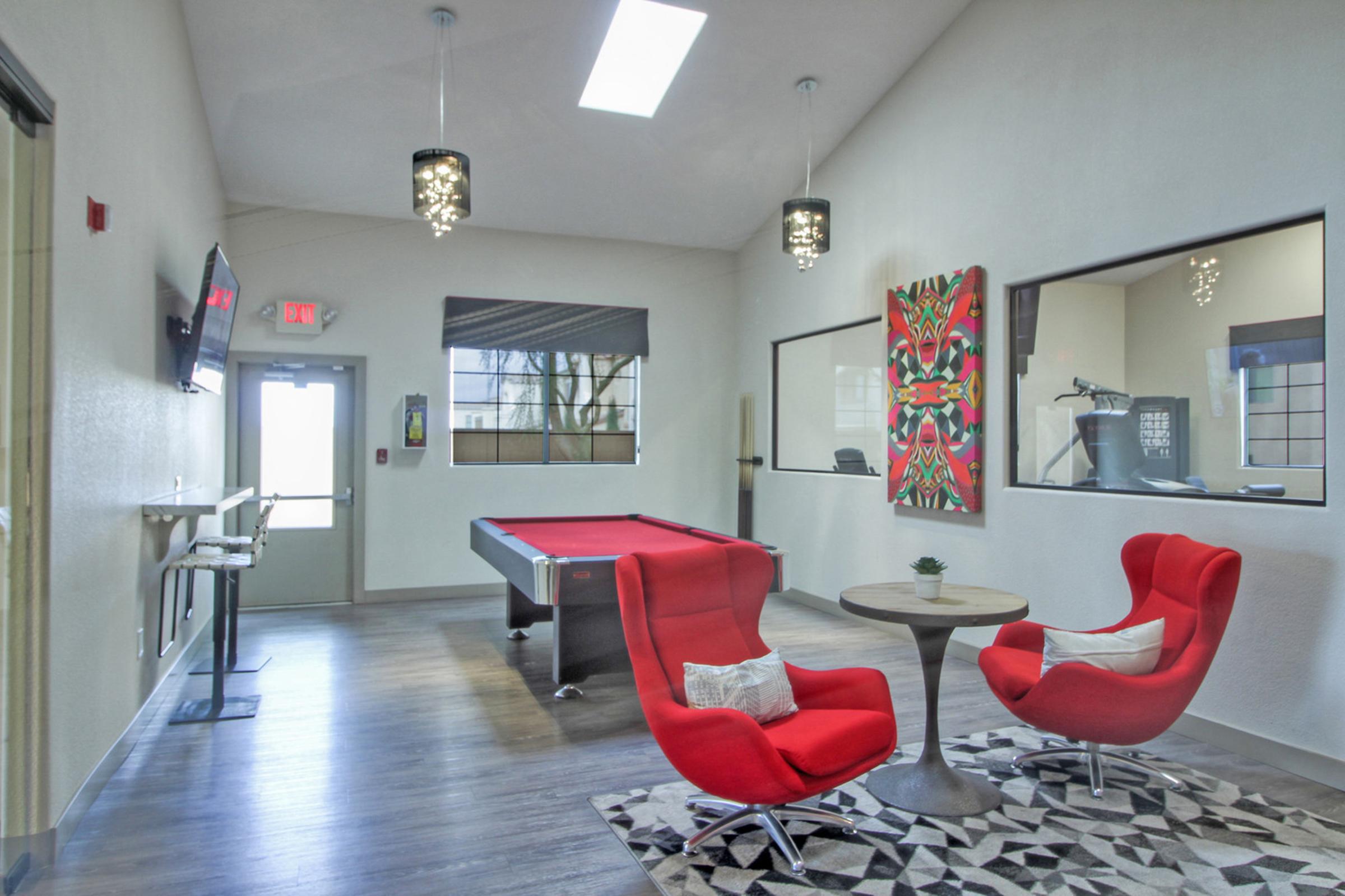 A bright, modern lounge area featuring two red armchairs around a small table, a pool table in the background, and large windows letting in natural light. The walls are adorned with artwork, and there are stylish light fixtures hanging from the ceiling, creating a welcoming atmosphere.